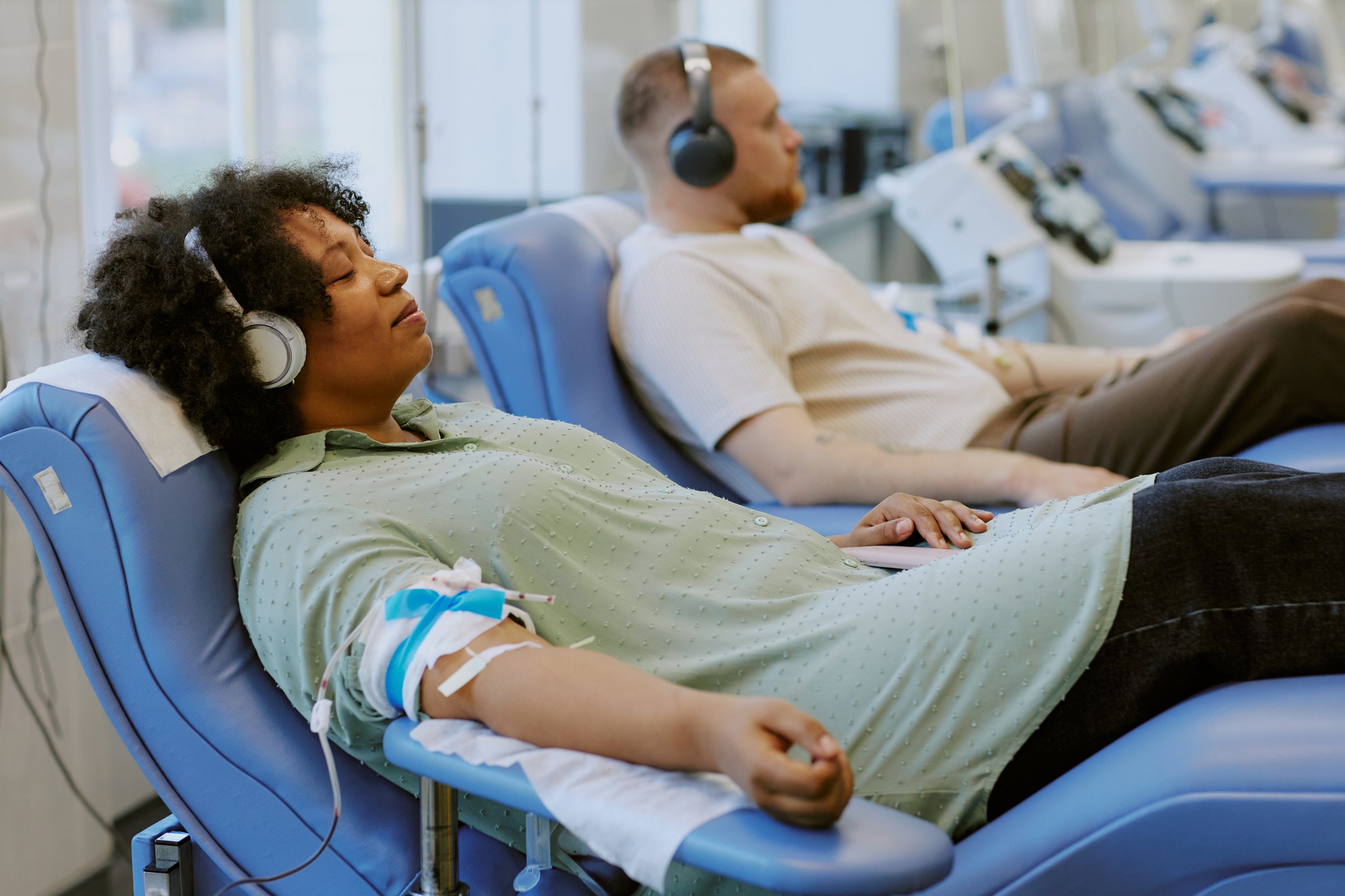 African American person donating blood while listening to music with other patients in background Donor sitting on a recliner with an IV in arm
