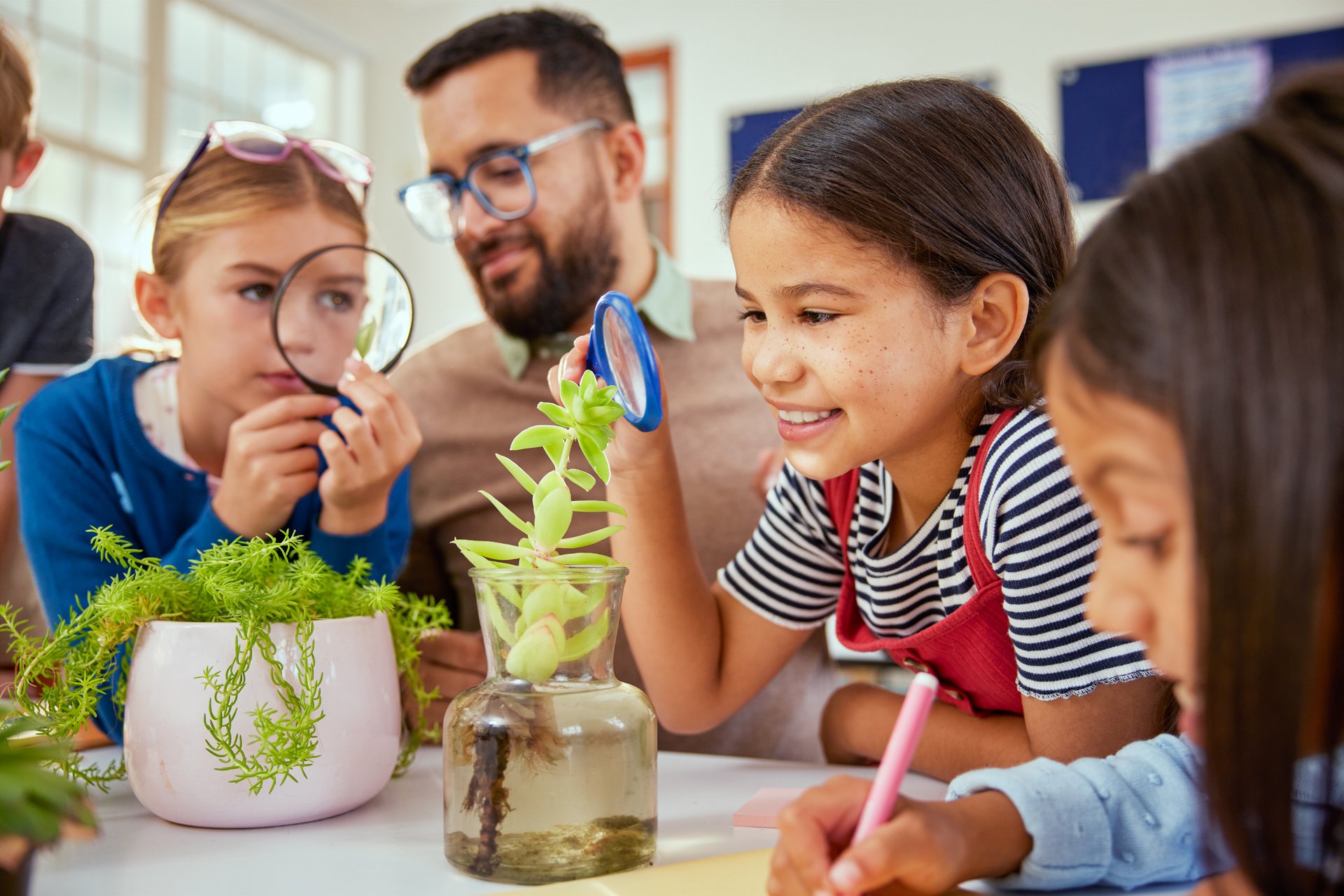 Happy schoolgirl exploring plants with magnifying glass in classroom. Teacher with multirthnic children in science class explaining plants biology. A group of schoolchildren sitting together and observing a plant with teacher.