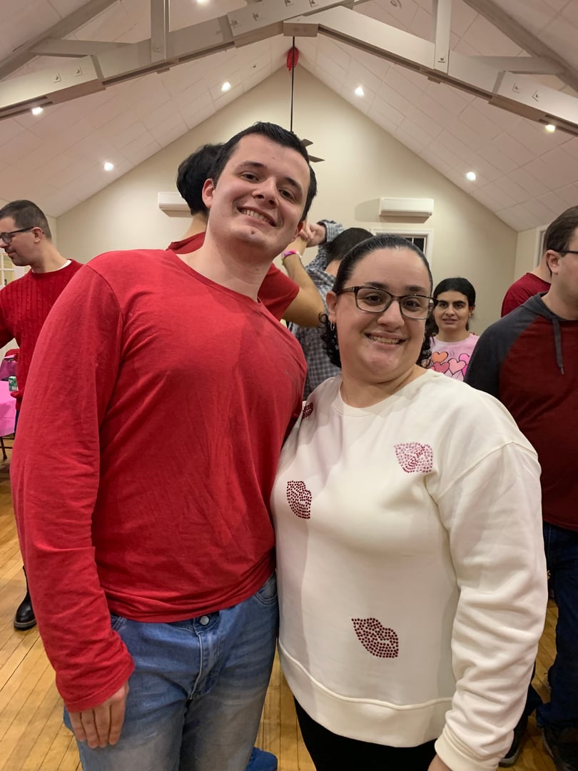 Two people smiling and posing indoors at an event, one in a red shirt, the other in a white sweater with red designs.