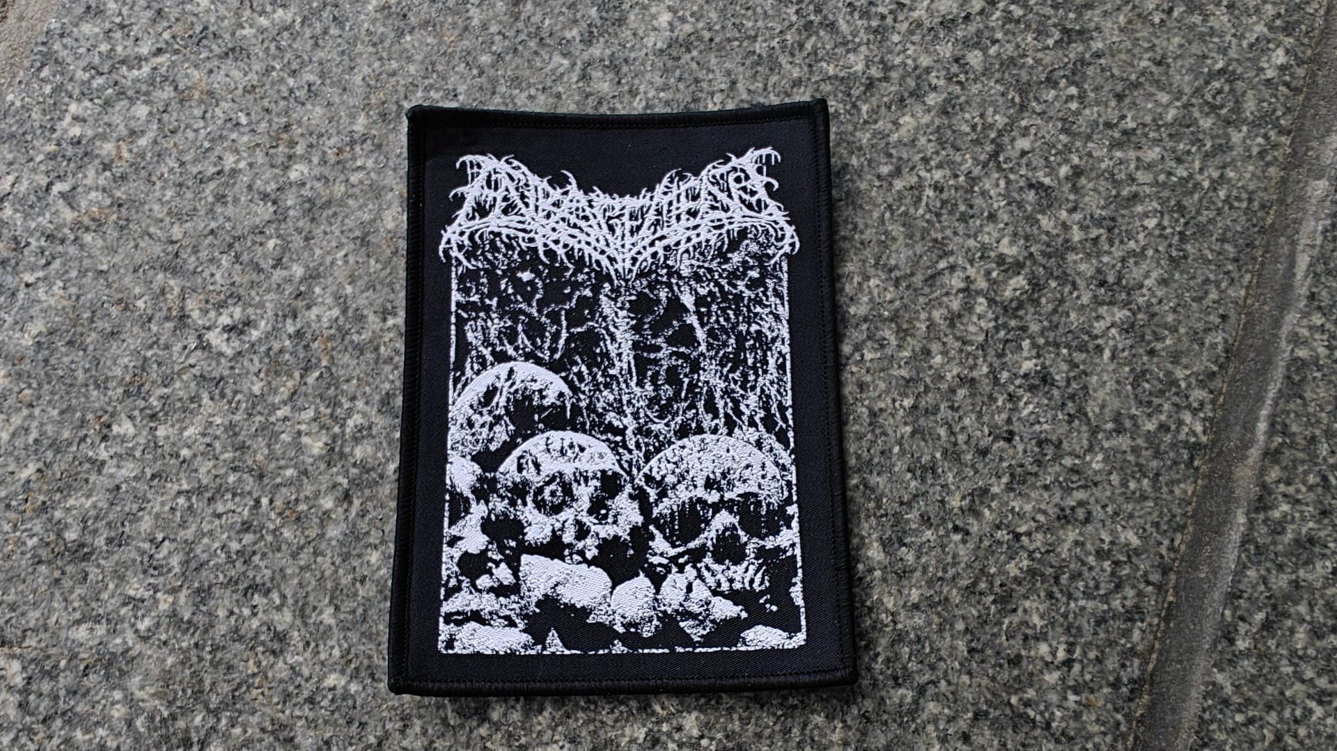 A rectangular patch on a stone surface features intricate white skull designs on a black background with gothic text.