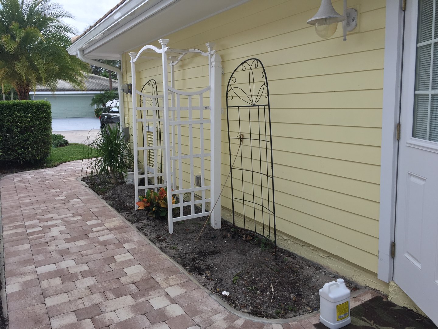A yellow house exterior with trellises, garden beds, and paver walkway. White door and decorative garden arches visible.