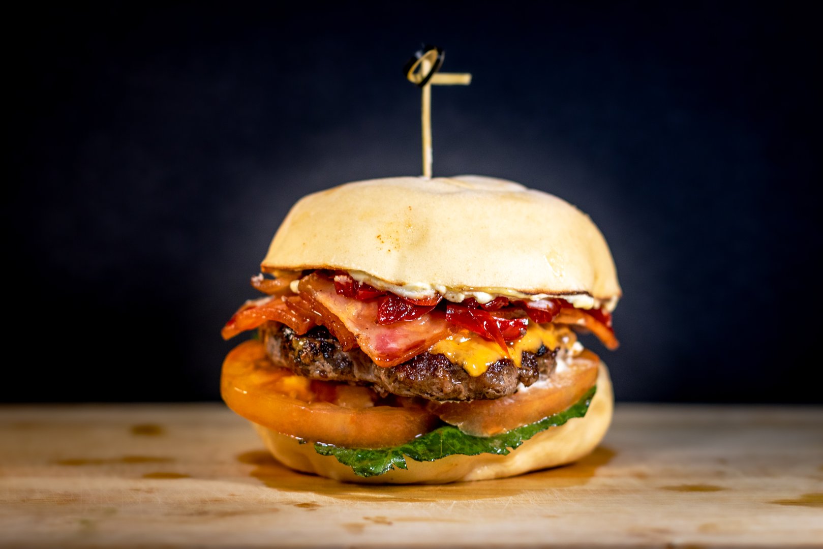 Close-up of a bacon cheeseburger with crispy bacon, cheddar cheese, tomato slices, lettuce, and sauce in a fresh bun, served on a wooden surface against a dark background.
