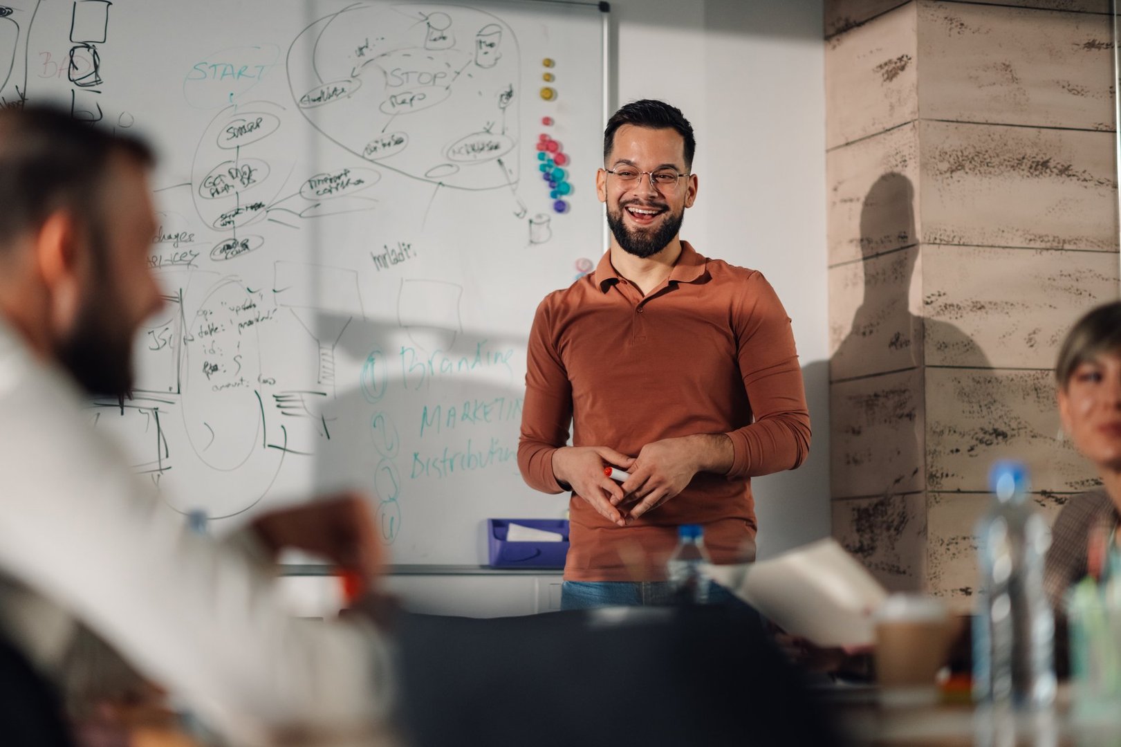 Smiling male entrepreneur leads a business meeting, presenting a project strategy on a whiteboard to his attentive colleagues in a modern office setting, fostering collaboration and innovation