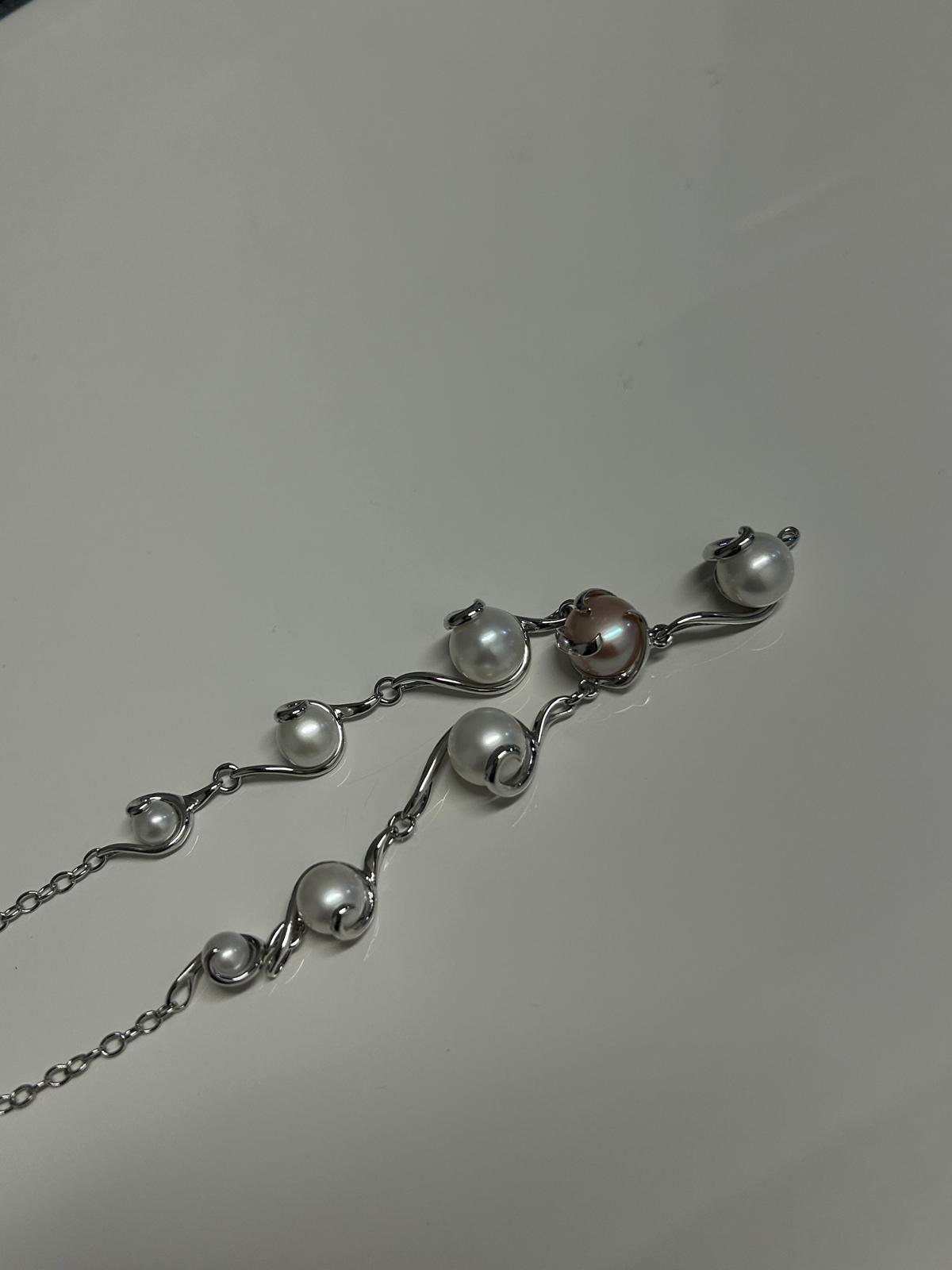 Silver necklace with multiple white and one brown pearl on a light background.