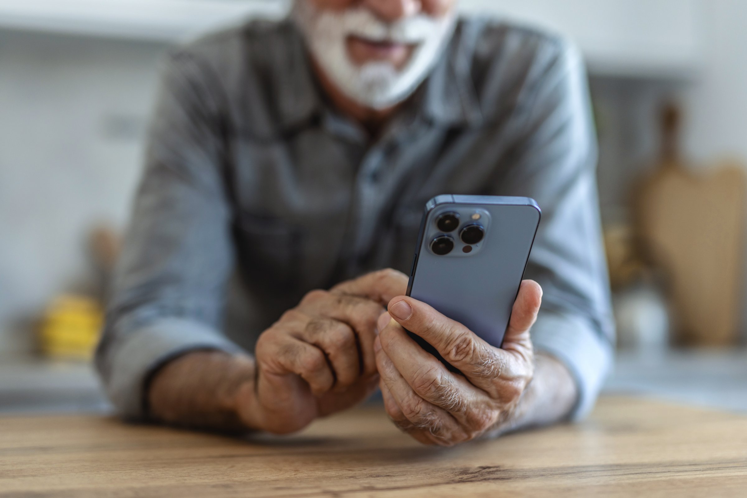 Mature man standing alone in kitchen, drinking orange juice and using smart phone. Elderly man, pensioner, sitting in kitchen and using mobile phone, he is in nursing home or in his apartment, concept of aging and happy retirement days.