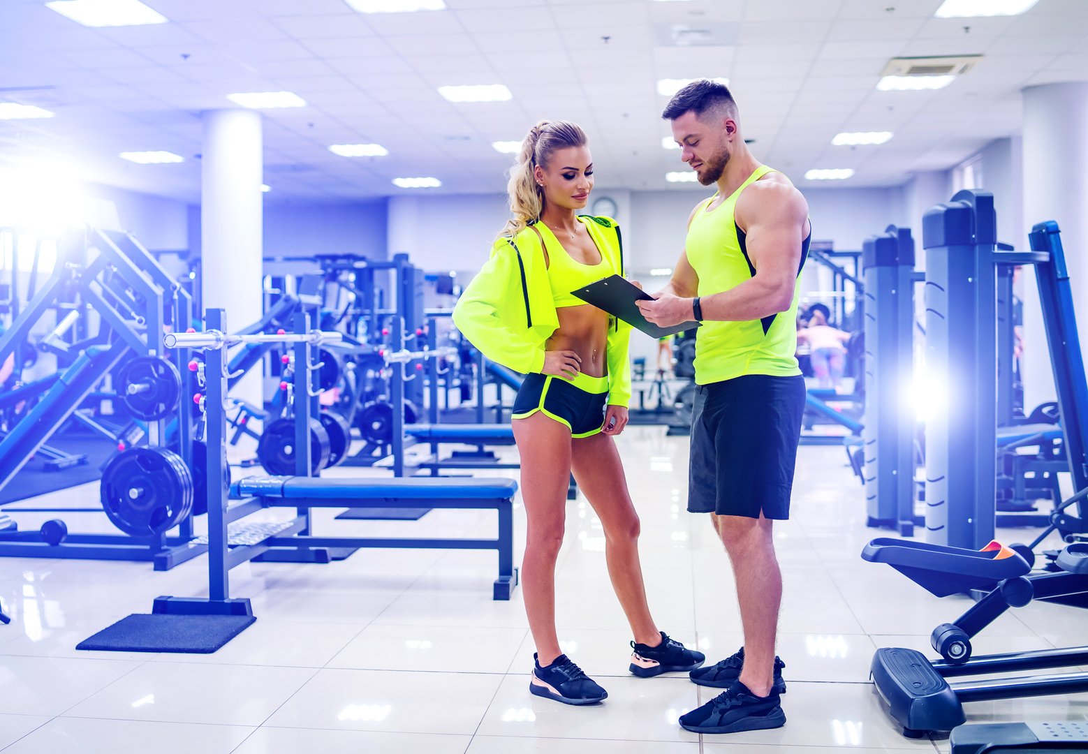 Personal trainer with a folder working with client woman at a gym. Creating a scedule. Wears bright clothes. The woman is in a jacket. Gym background.