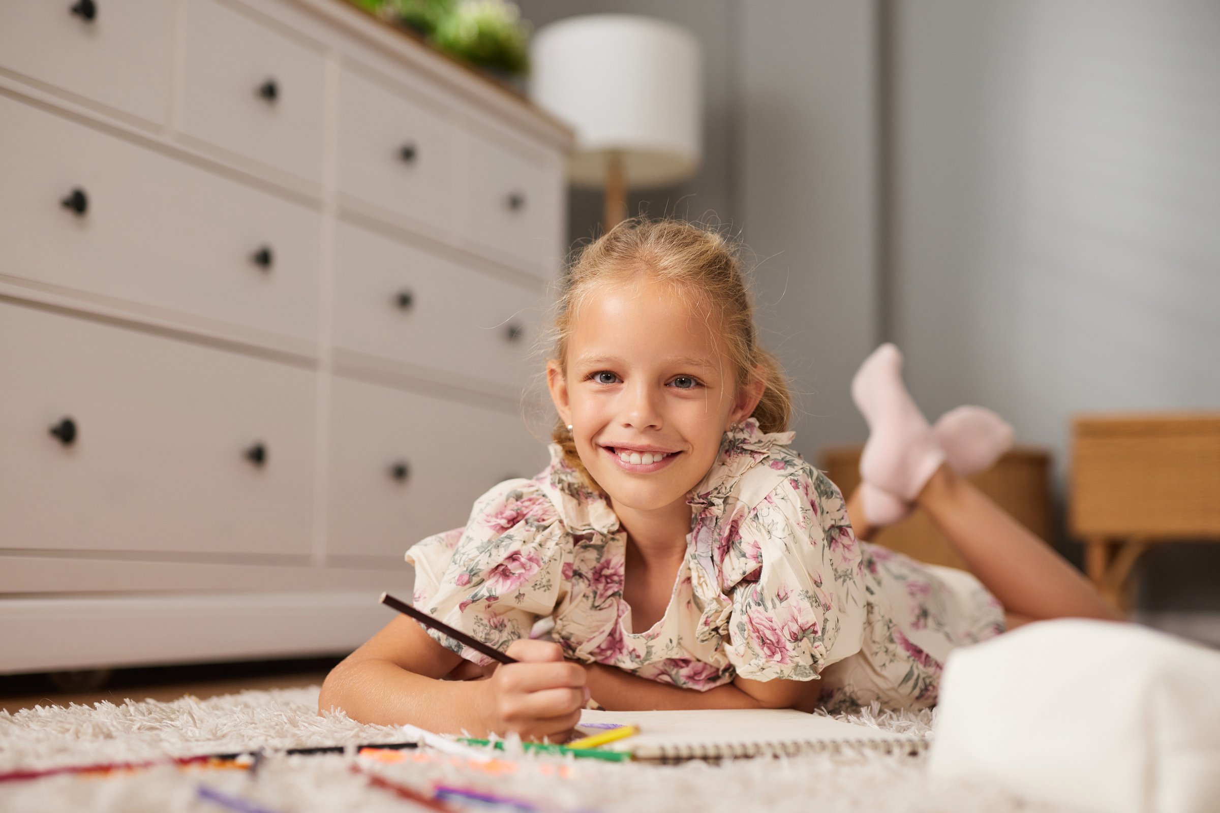 A bright young girl lies on her stomach on a plush rug in a warm living room, happily drawing with colorful crayons on a large sheet of paper while smiling.