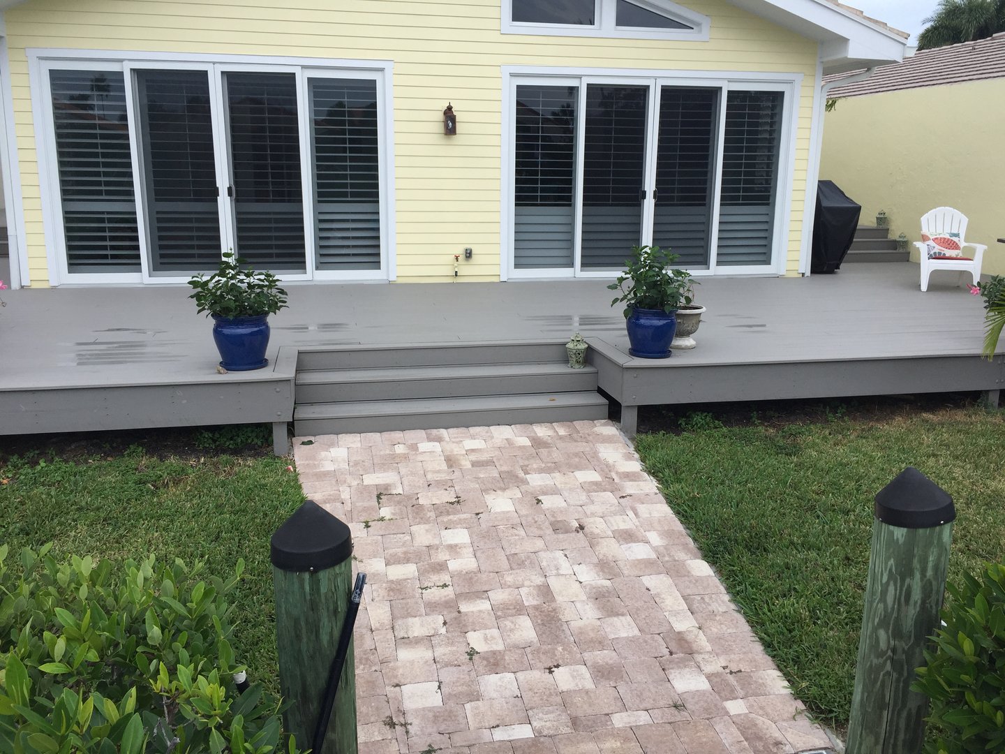 Backyard deck of a yellow house with sliding glass doors, gray steps, potted plants, and a brick walkway.