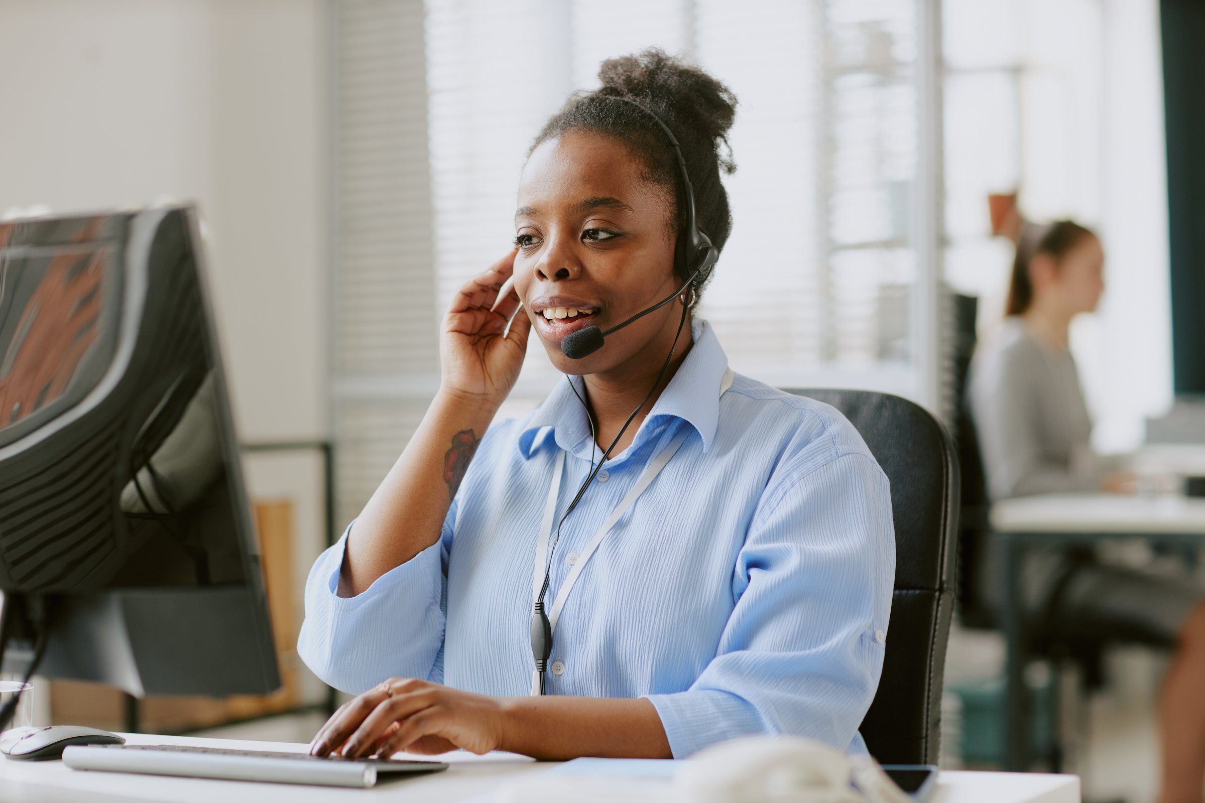 Young African American female worker dressed in pastel blue blouse helping client remotely via headphones while looking at details on monitor