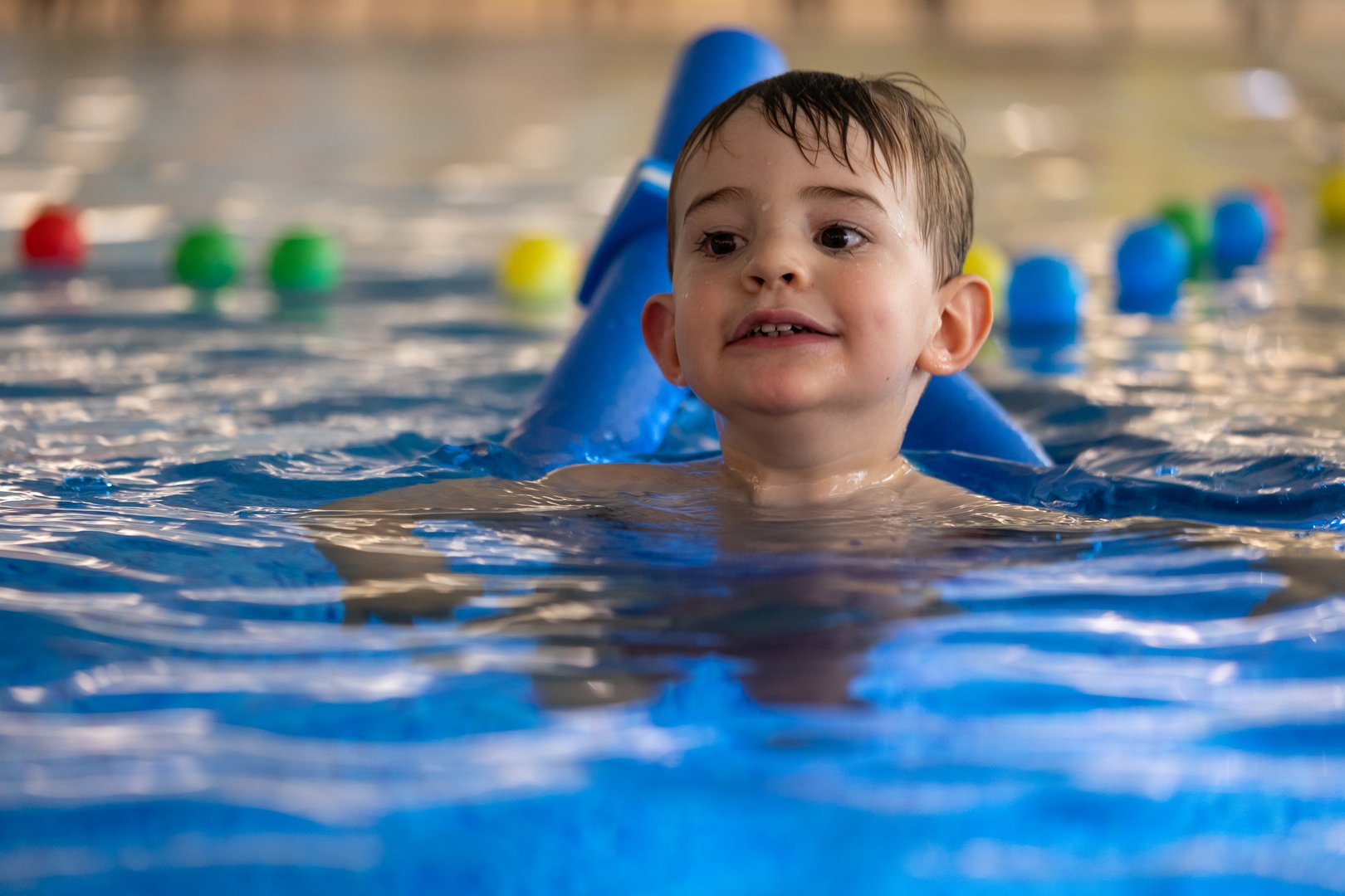 Young boy enjoying his swimming lesson, using a blue floatation device in an indoor pool, promoting water safety and early childhood development