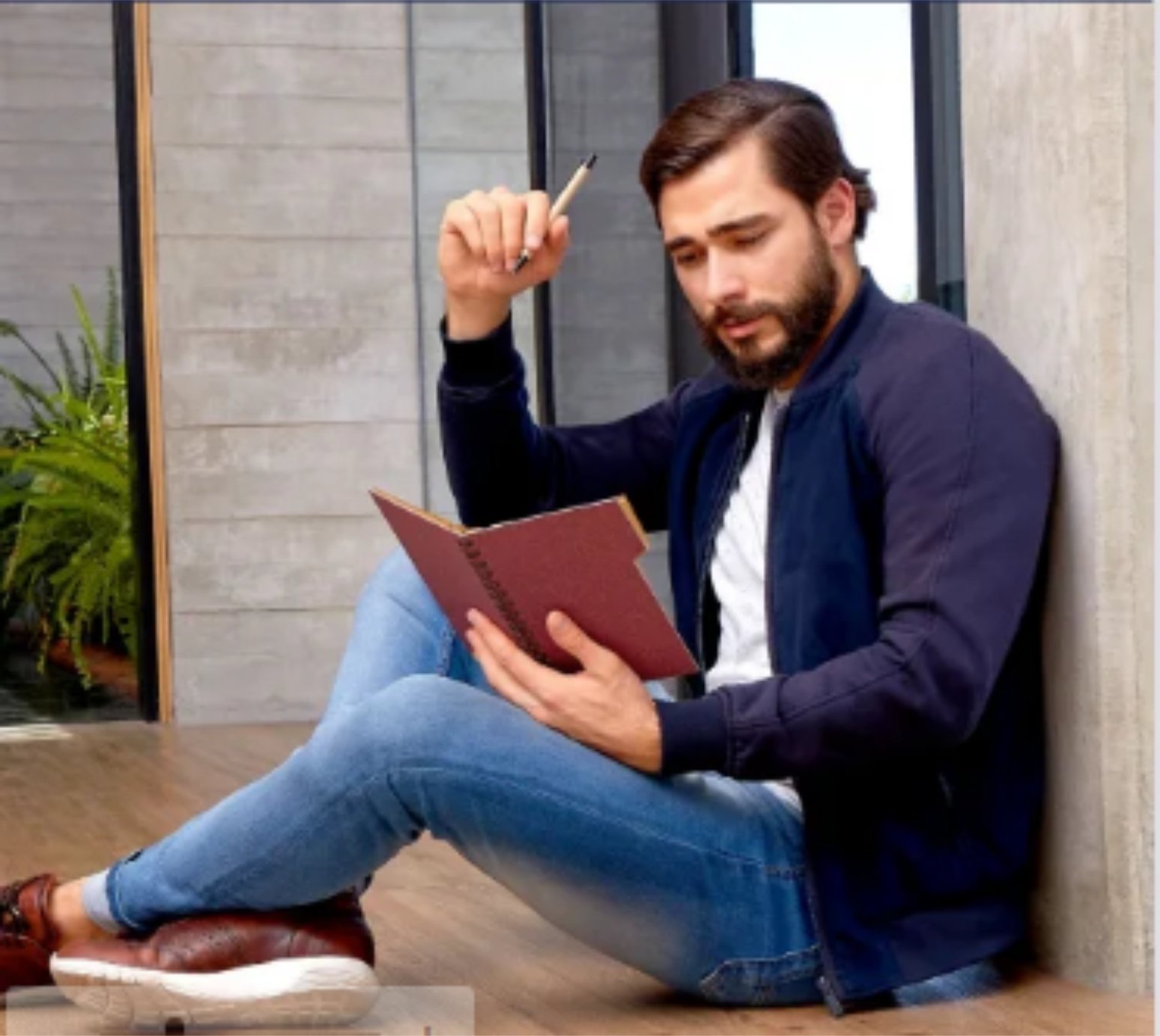 Man sitting on floor with a notebook and pen, leaning against a concrete wall, dressed in casual attire.