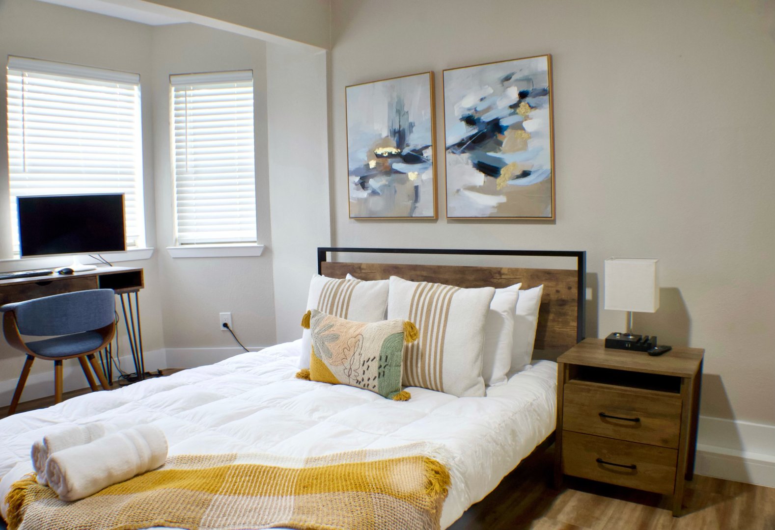 Modern bedroom with a wooden bed, abstract art, desk with computer, and window light. Decor includes pillows and a throw.
