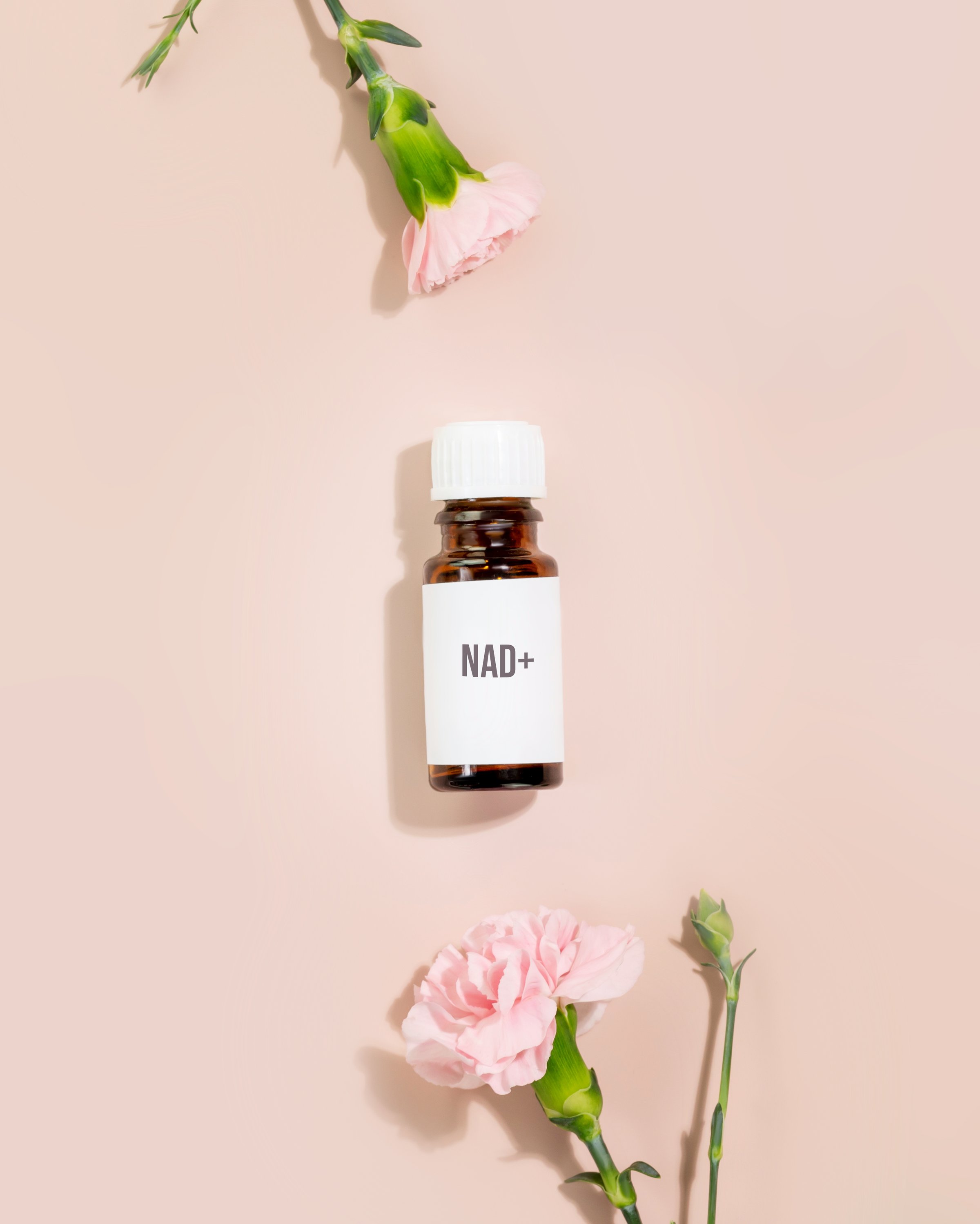 NAD essence in vial with white cap surrounded by pink carnations on nude pastel background. Concept of NAD beauty products, cellular beauty, anti-aging trends, and modern wellness products