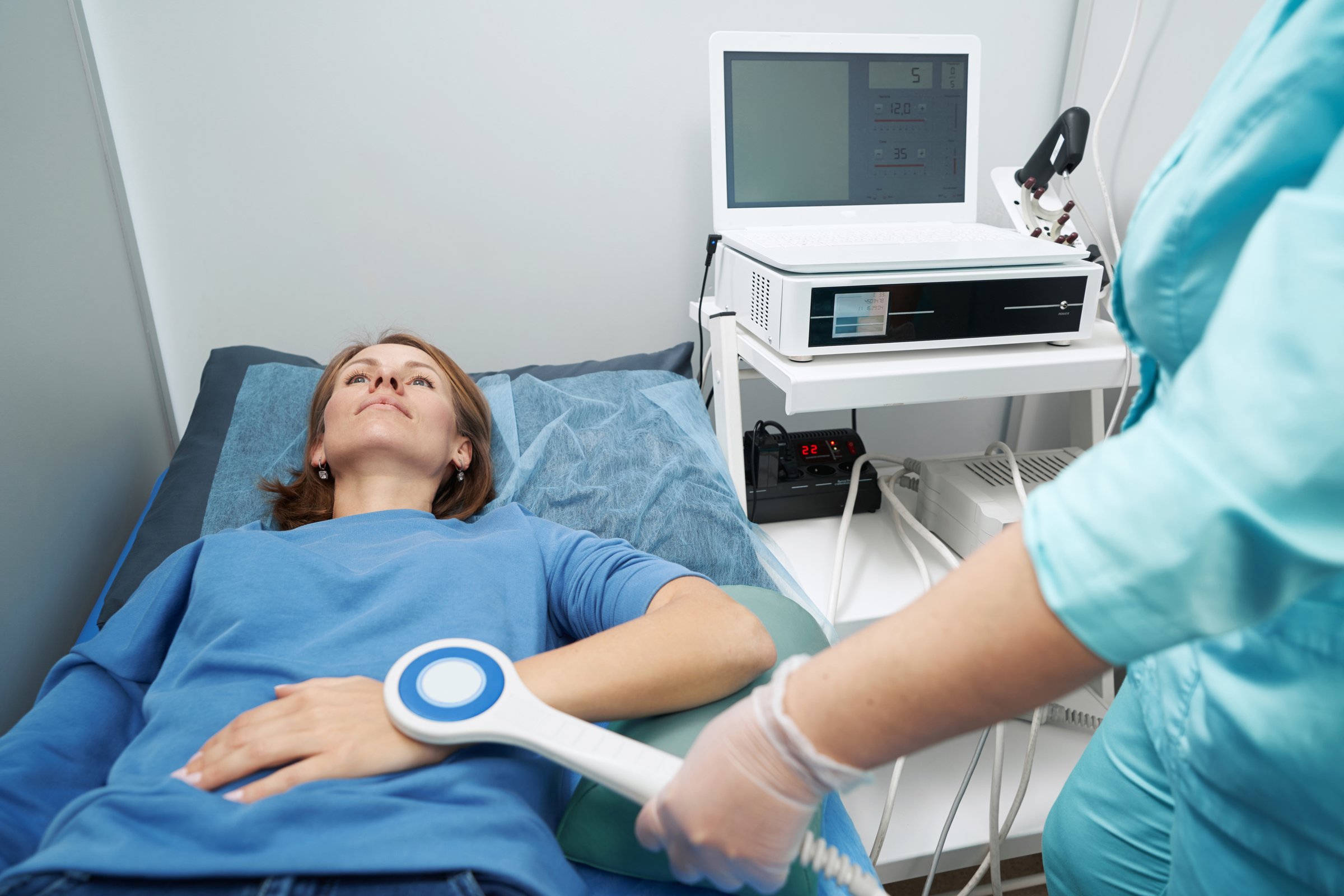 Female patient lying on hospital daybed while doctor performing physiotherapy procedure on woman arm with medical equipment