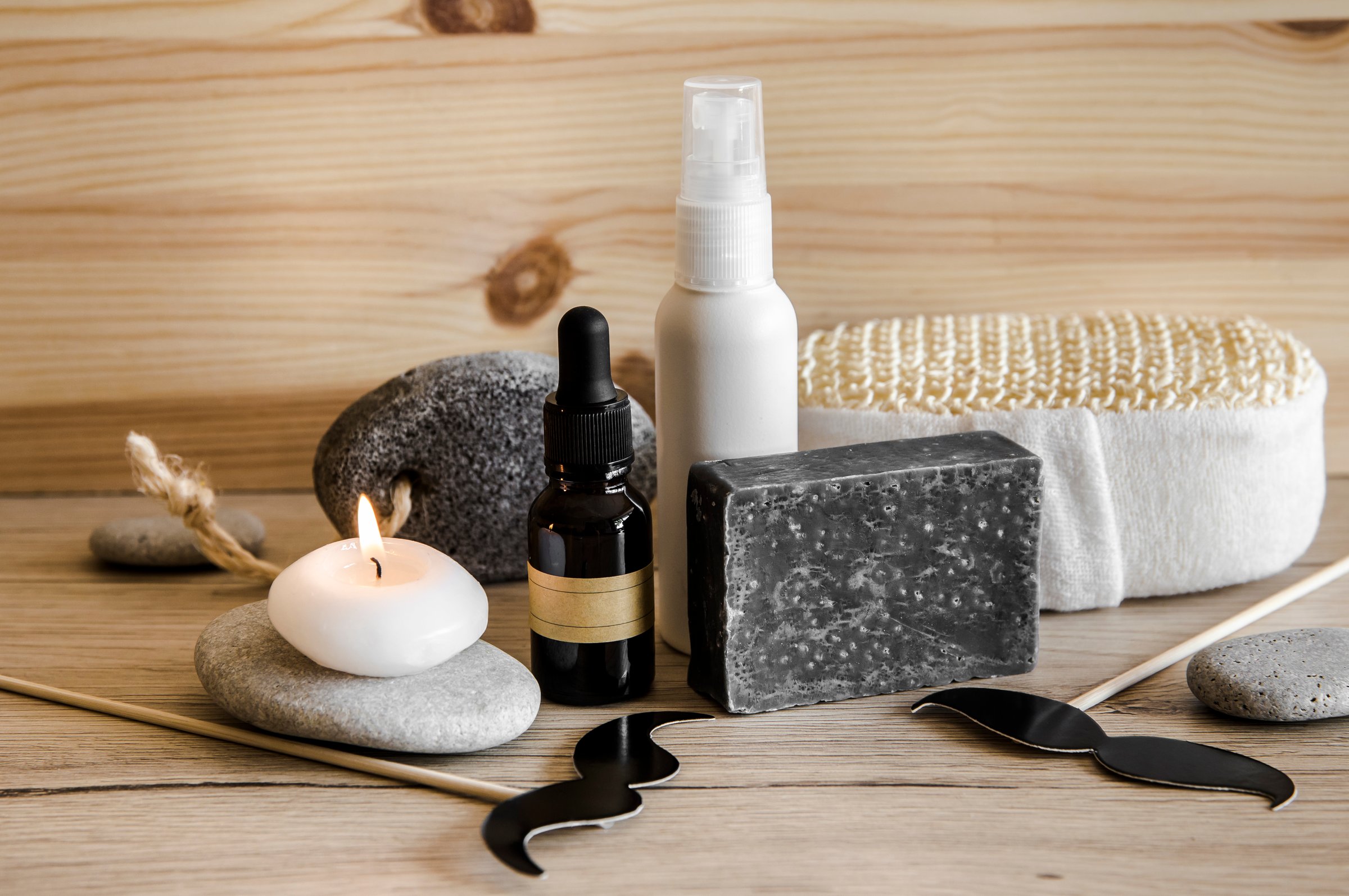 Above view set of various man spa products things. Natural wood disc background with flat sea stones. Gray clay soap bar, beard oil, moisturizing cream, candle burning. Funny mustache shape figurines.