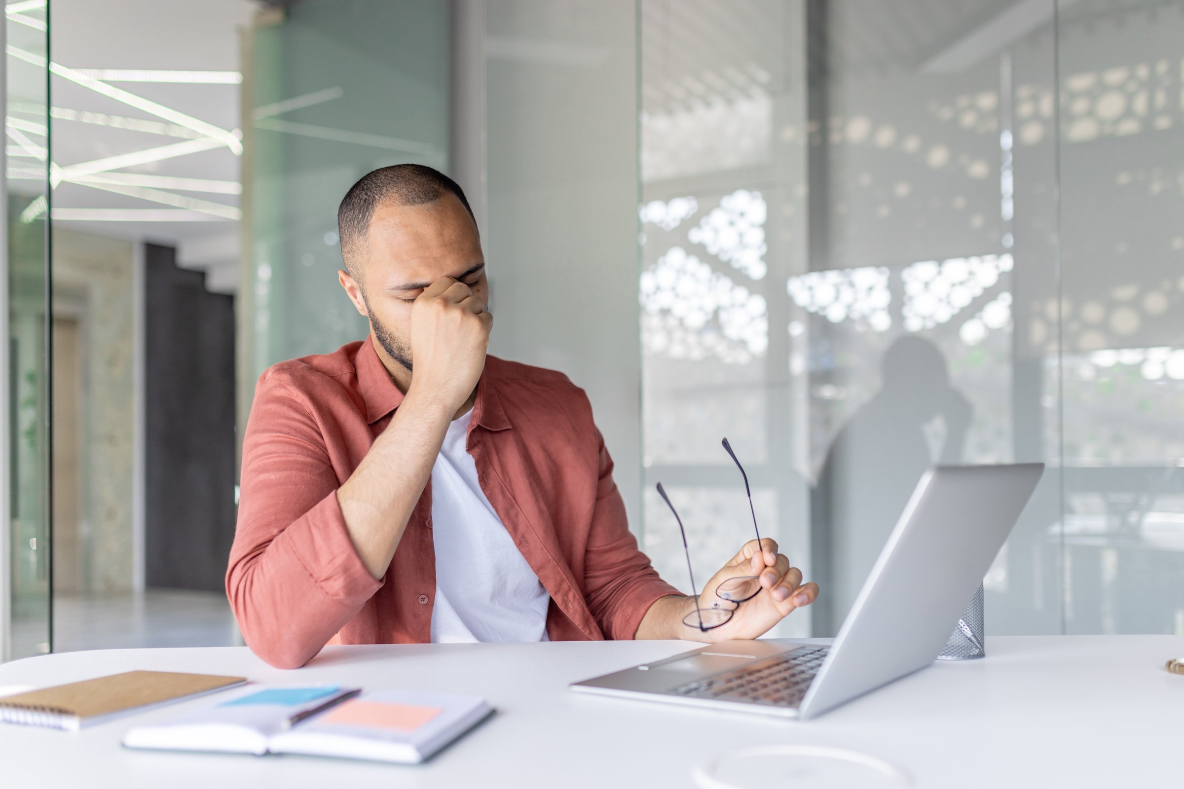 Man has severe headache and dizziness at workplace inside office, office worker working sick with laptop at workplace, suffering from eye pain.