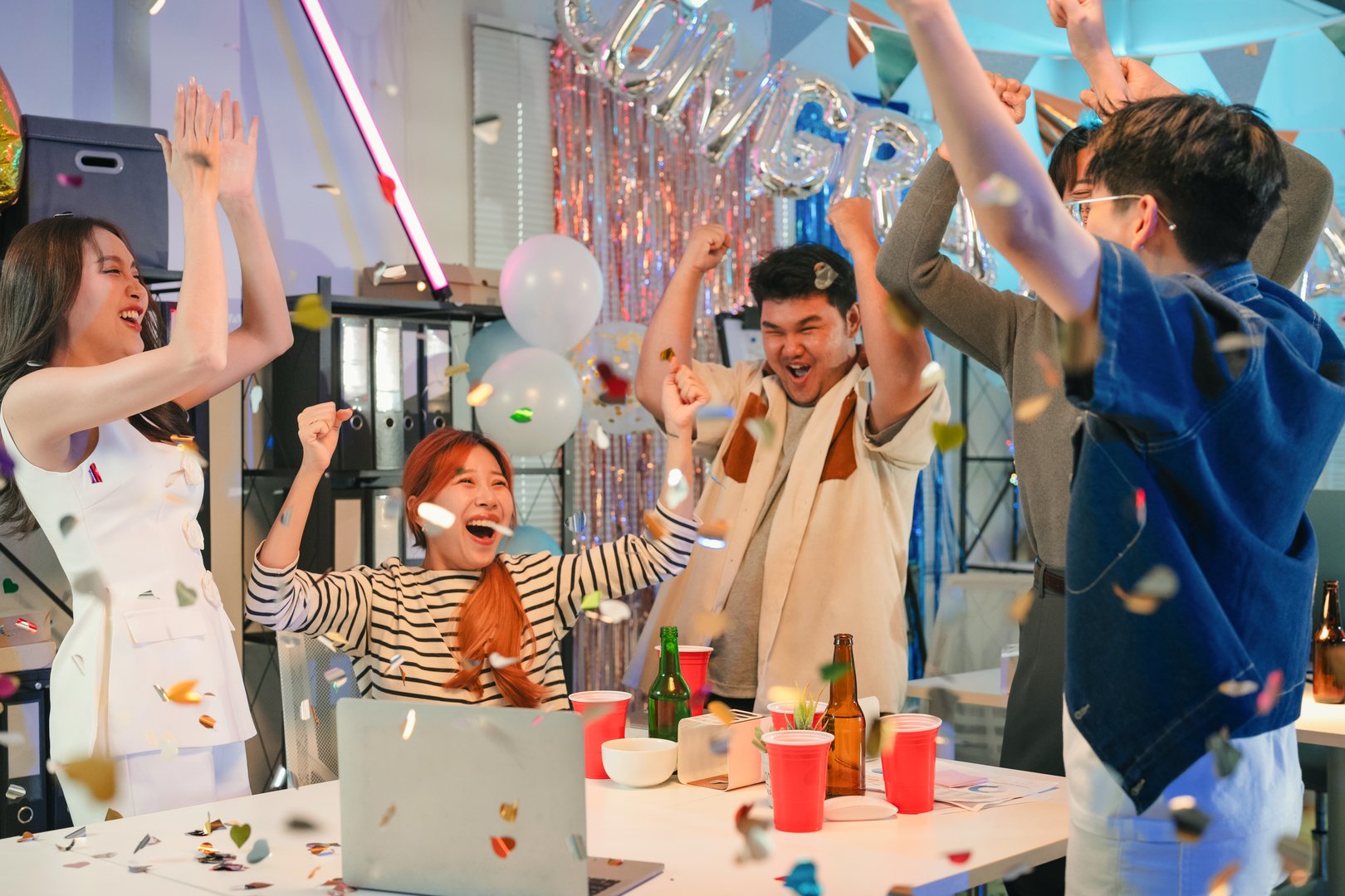 Excited office team celebrates incredible profit growth and victory results announcement together, cheering enthusiastically while monitoring financial performance on screen during triumphant nighttime celebration at workplace