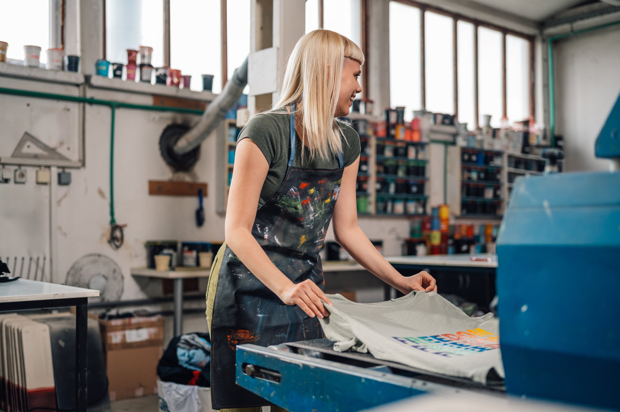 Smiling female printing workshop worker putting silkscreen printed t-shirt into drying machine at facility. Female graphic technician finishing process at printing shop. Females in printing industry