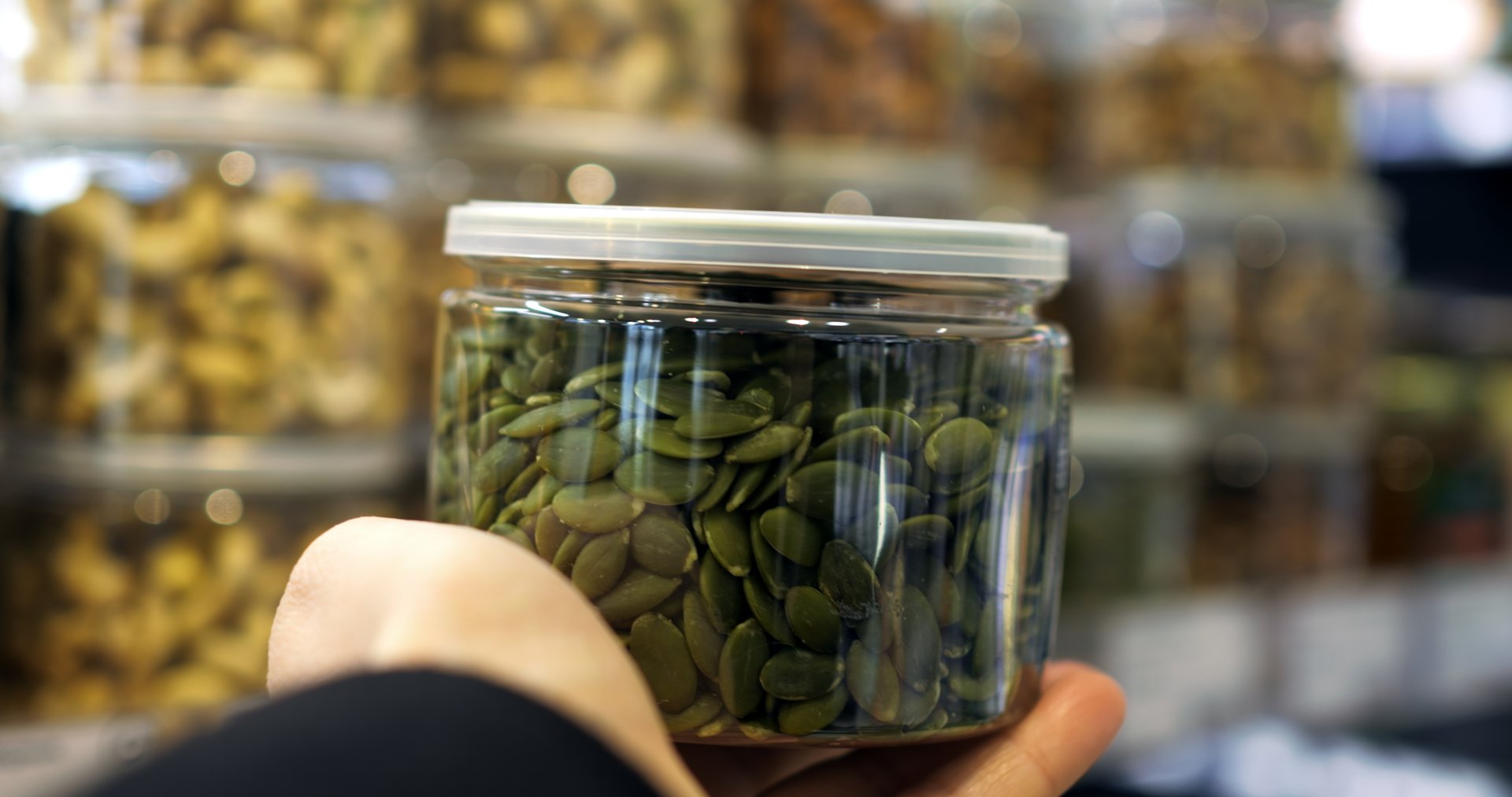 Customer hand is holding a transparent plastic jar full of green pumpkin seeds, with a blurred background of other nuts and dried fruits in similar containers