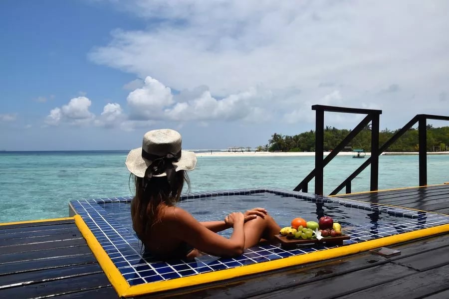 Person in a sunhat relaxes in an overwater pool with fresh fruit on the side, overlooking a tropical island view.