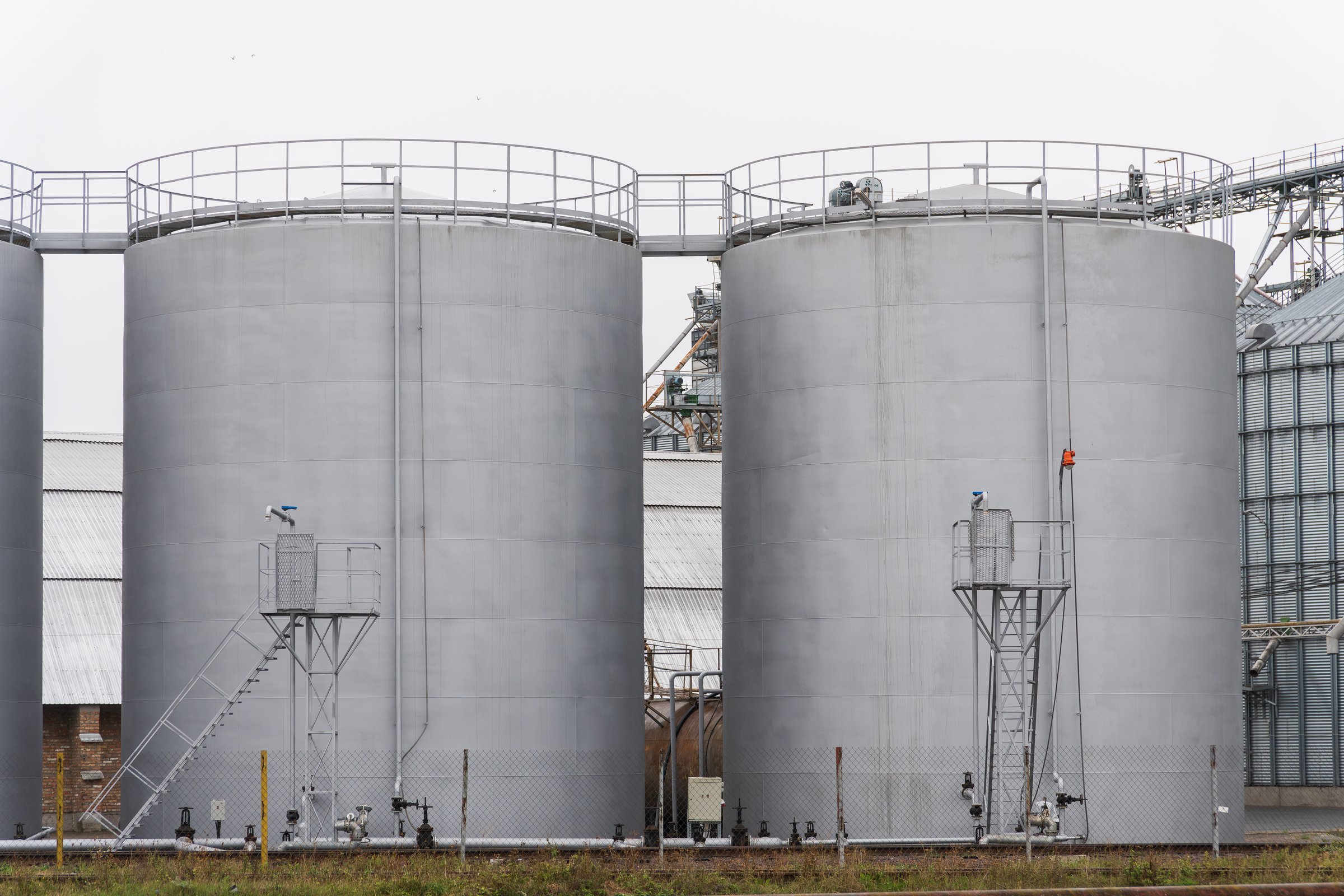 Two large steel storage tanks are positioned side by side at a chemical facility. Safety equipment is evident, indicating an industrial setting under cloudy conditions.