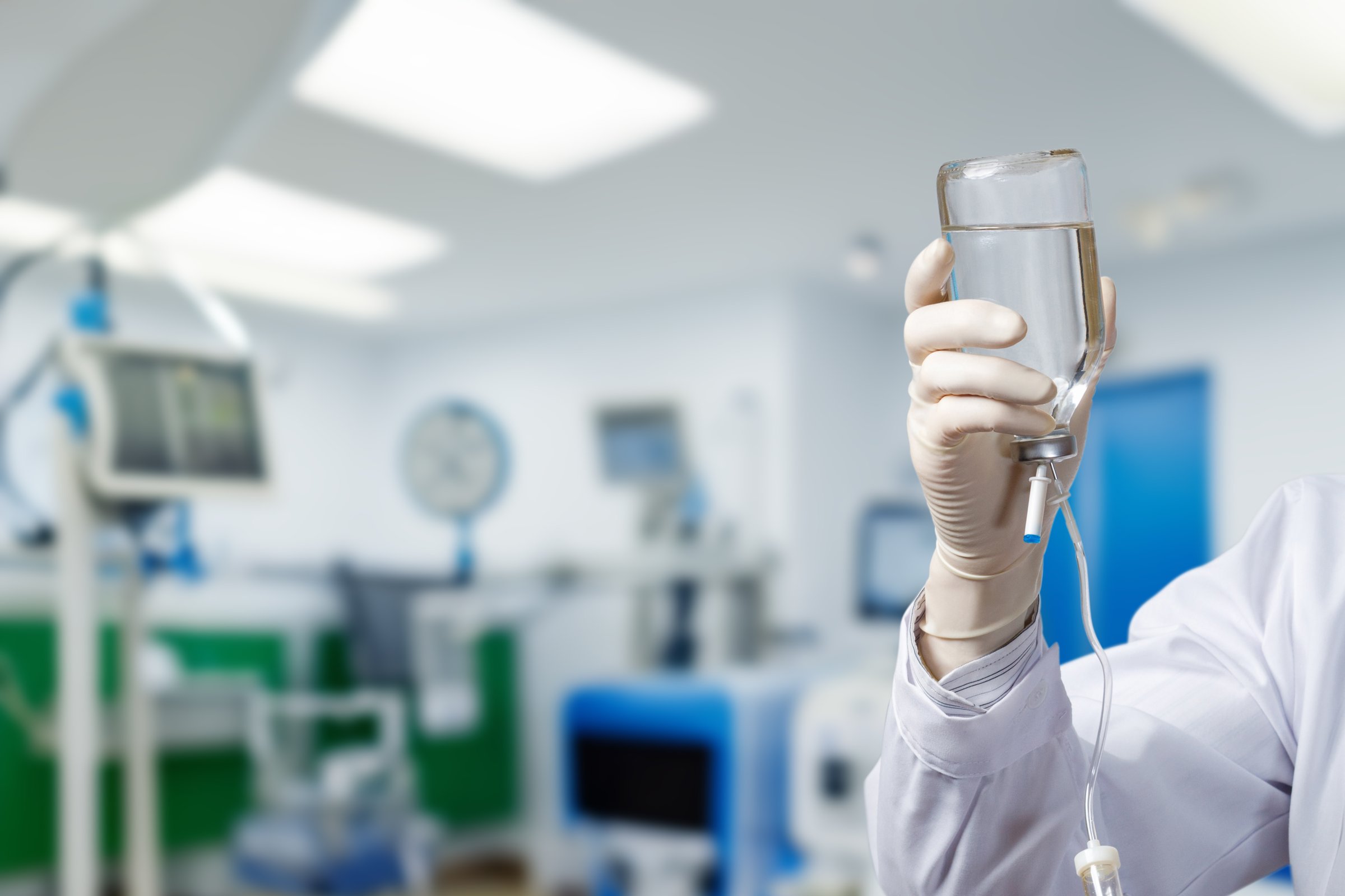 Holding a medical drip in a healthcare facility. A hand in a medical glove holds a clear drip bag while equipment fills the background.