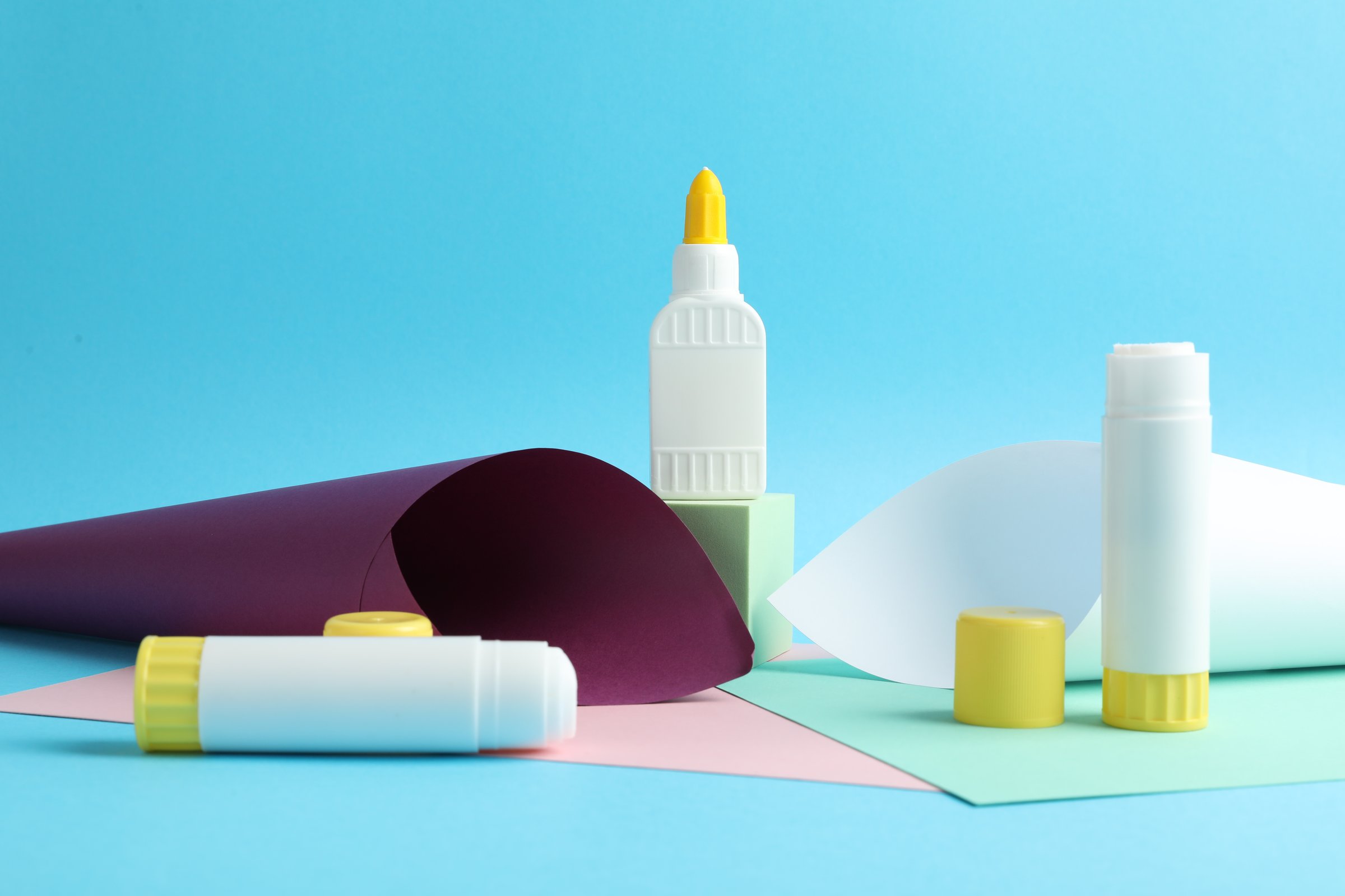 Glue in bottle, sticks and color paper on light blue background, closeup