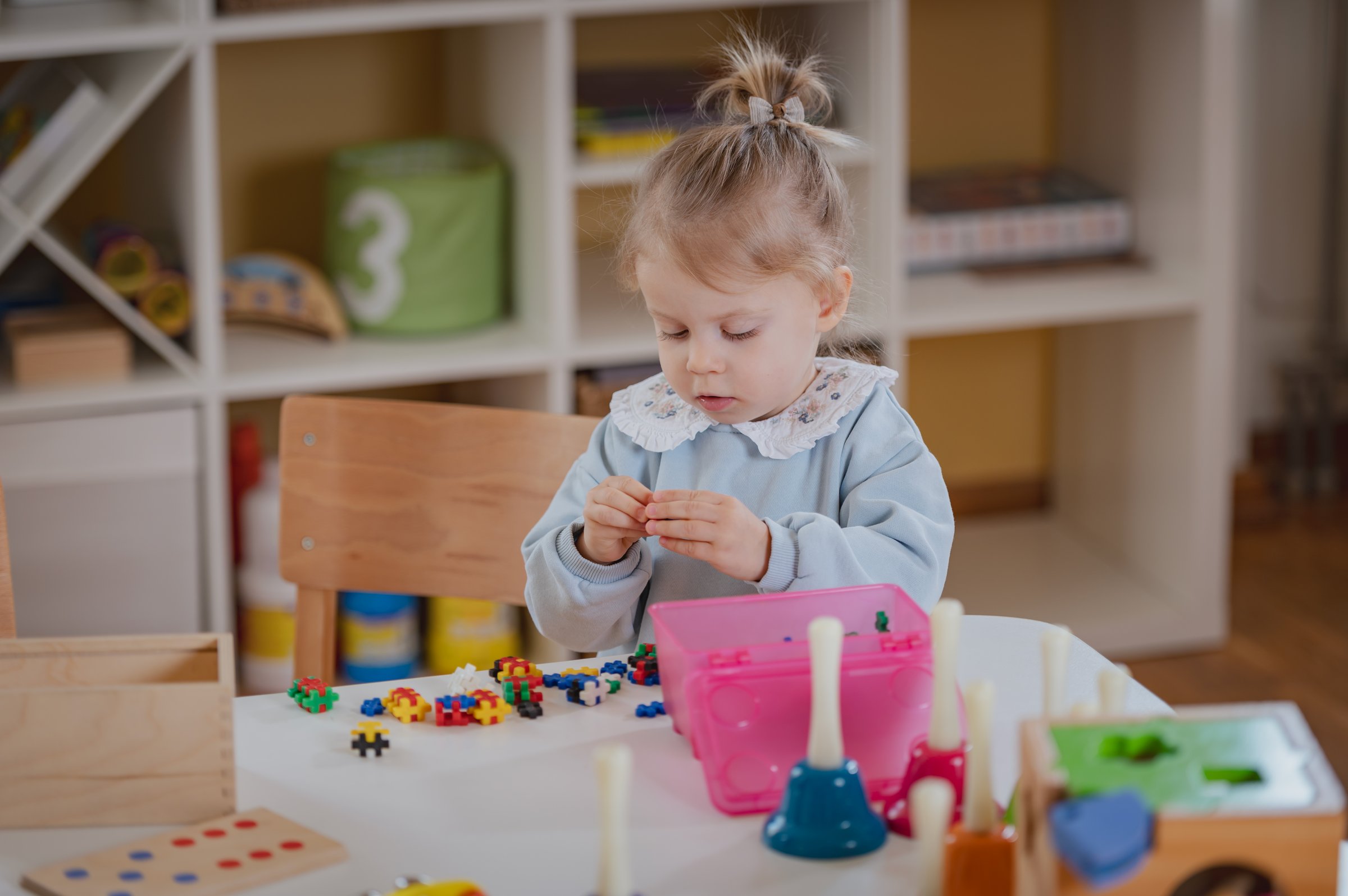 A young girl with blonde hair in a bun, wearing a light blue sweater, sits at a table in a preschool, playing with colorful beads and toys, focused on her activity