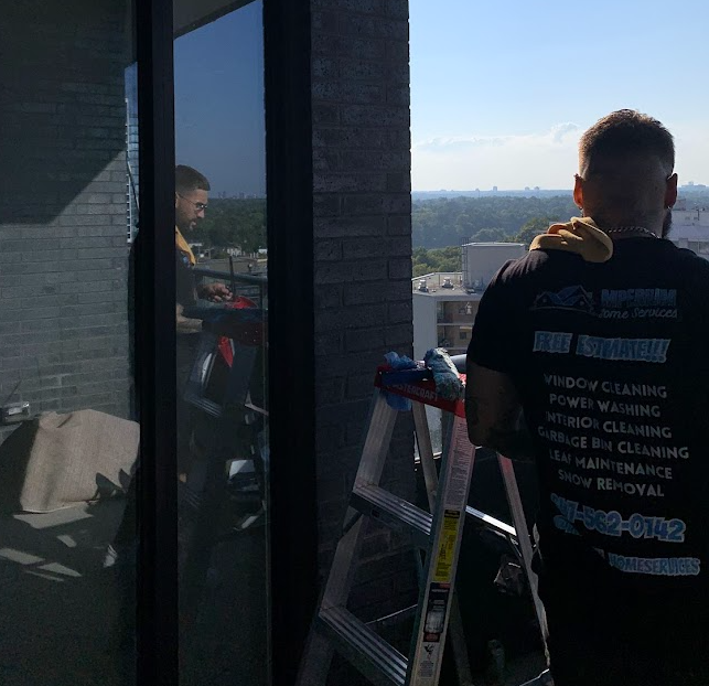 Two workers clean windows on a balcony, one standing on a ladder, with cityscape visible in the background.