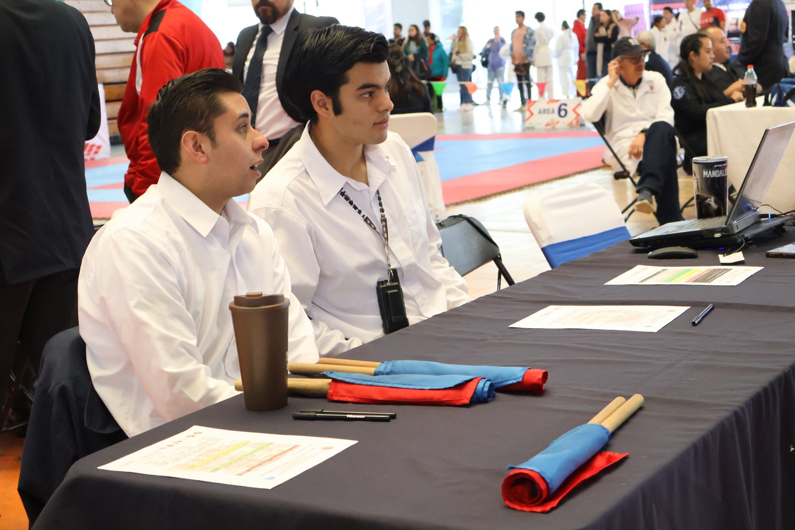 Two men sit at a table with papers and colored flags at a sports event, surrounded by people in martial arts attire.