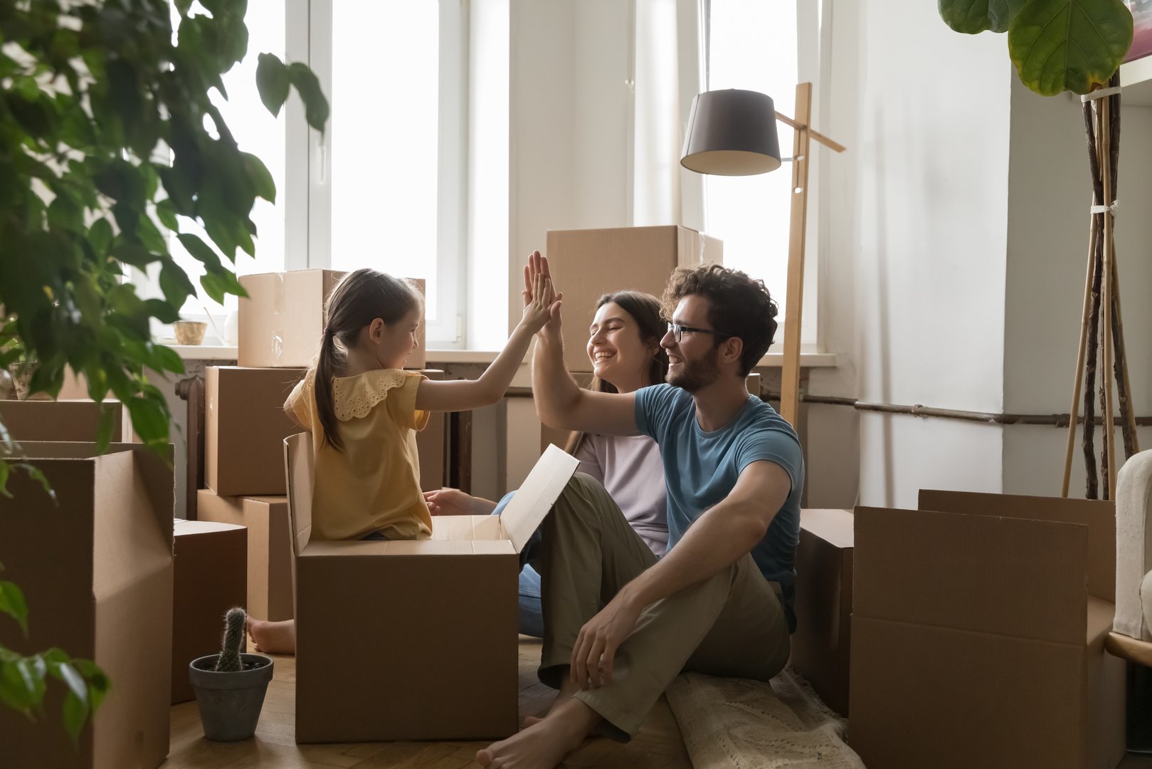Young family with little daughter celebrate relocation day, feel happy, giving high five gesture seated on floor near heap of cardboard boxes. Bank loan, tenancy, first dwelling, housing improvement