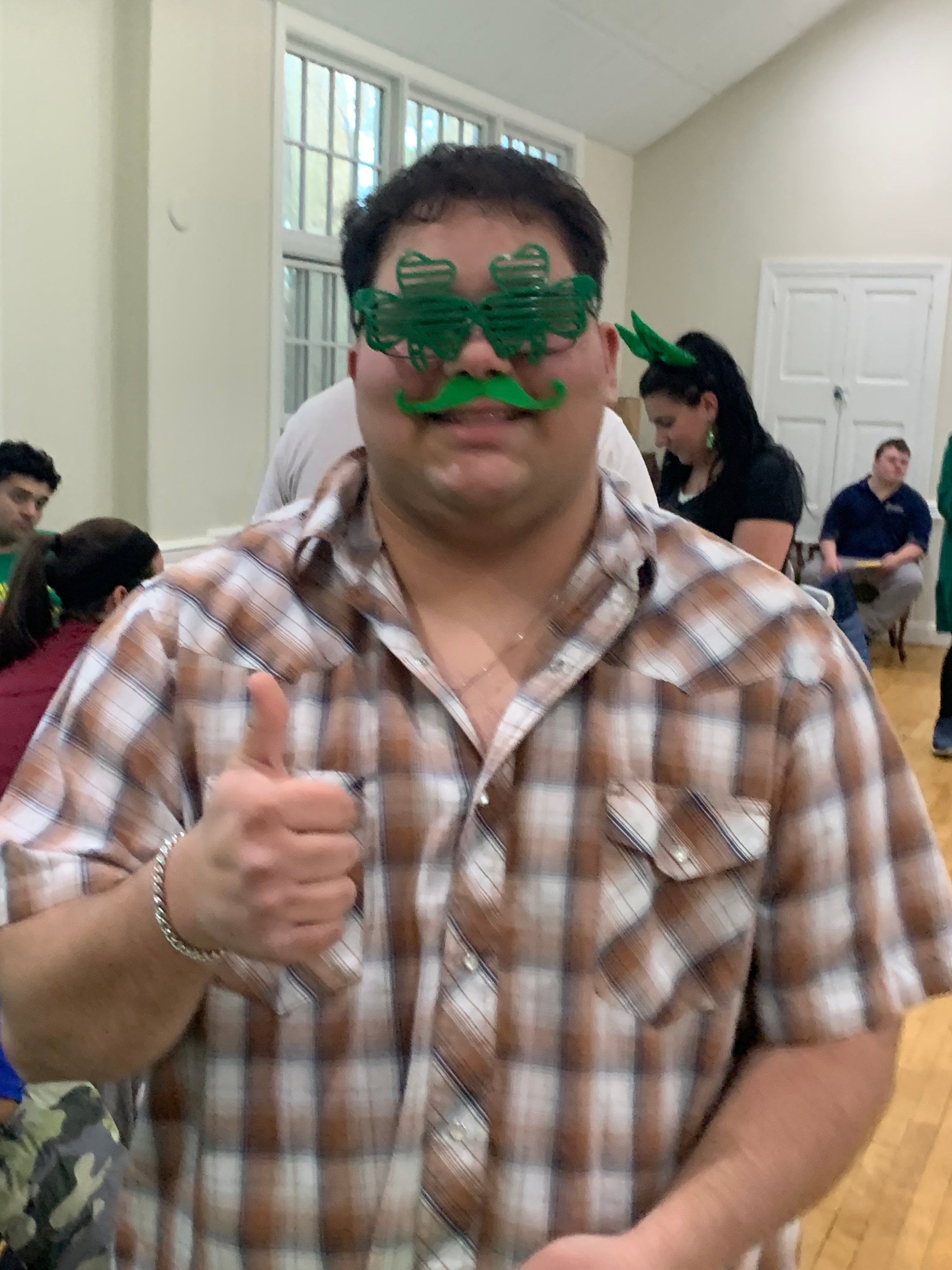 Person wearing green novelty glasses, mustache, and a plaid shirt, giving a thumbs-up in a room with people.