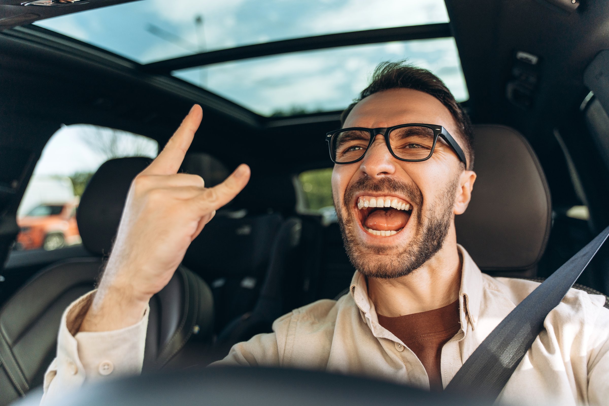 Portrait of overjoyed, happy middle aged man wearing casual clothes and stylish eyeglasses, driving new car, showing rock n roll gesture, looking at camera. Transportation concept