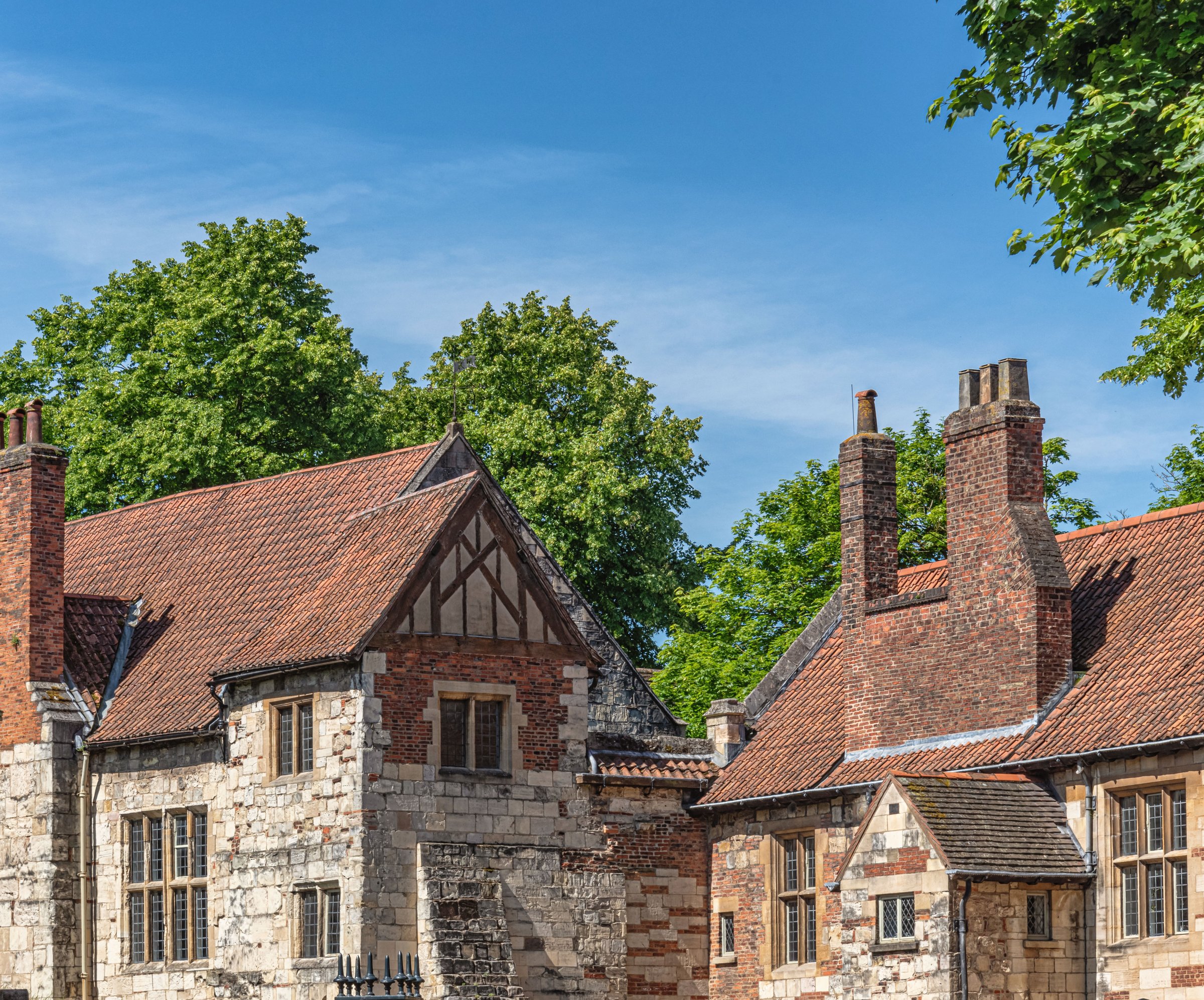 York, UK. June 2, 2024.  Two ancient buildings, a part of Kings Manor in York, stand in the sunshine surrounded by trees. A sky with cloud is above.