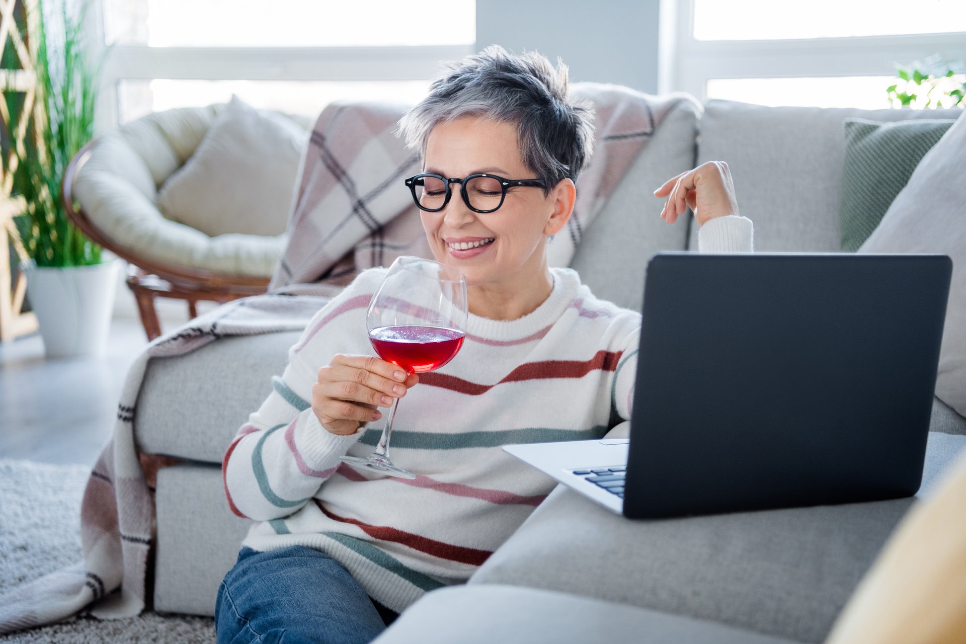 Photo of cheerful peaceful grandmother hold wine glass watch movie laptop spend pastime modern flat inside.