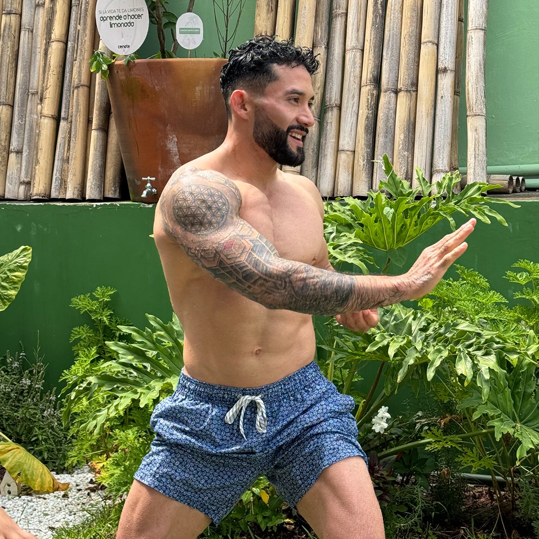 Man with tattoos performing martial arts stance in a garden, wearing blue shorts, surrounded by plants and bamboo.