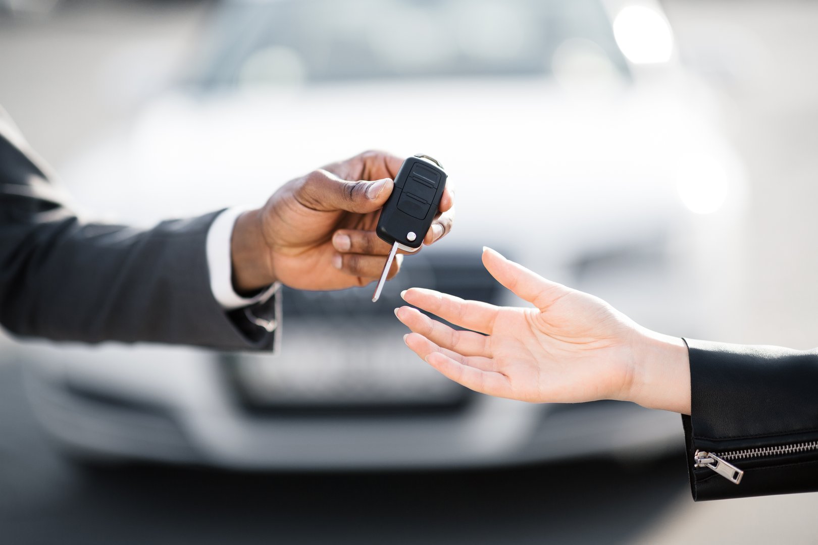 A person in a suit extends a car key to another individual, who is reaching out to receive it. The background features a parked vehicle under clear skies, indicating daytime activity.