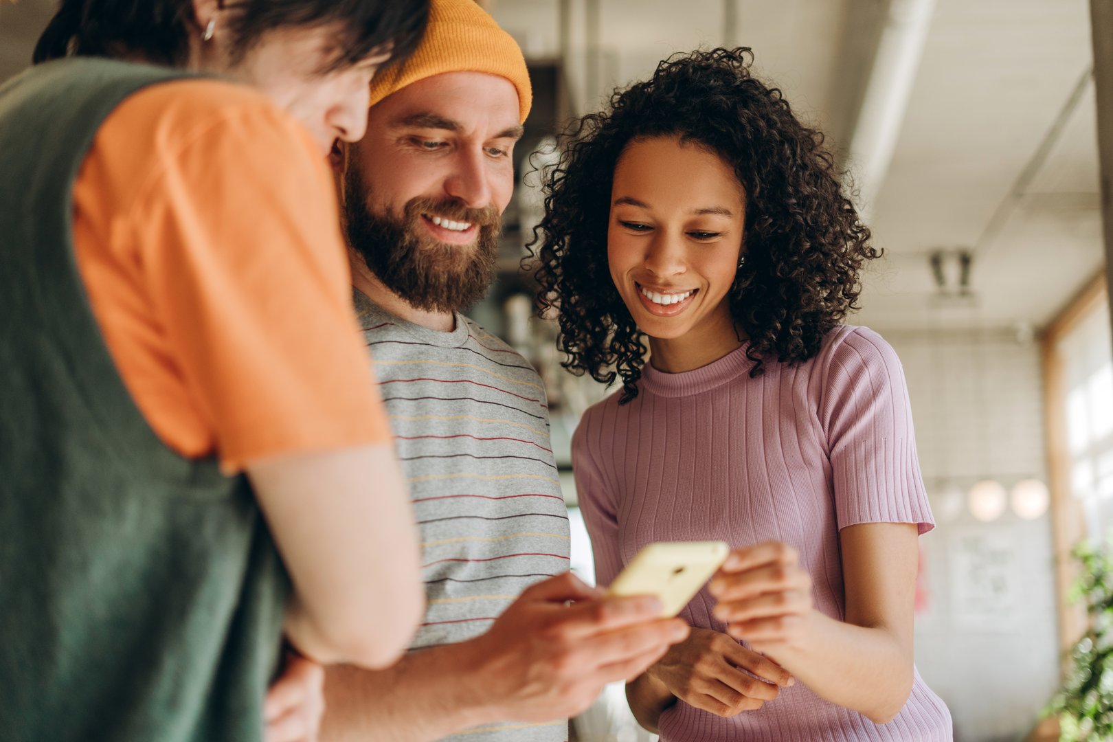 Three young colleagues smiling and collaborating on a project, sharing ideas while looking at a smartphone in a modern office filled with creativity and innovation. Online technology concept