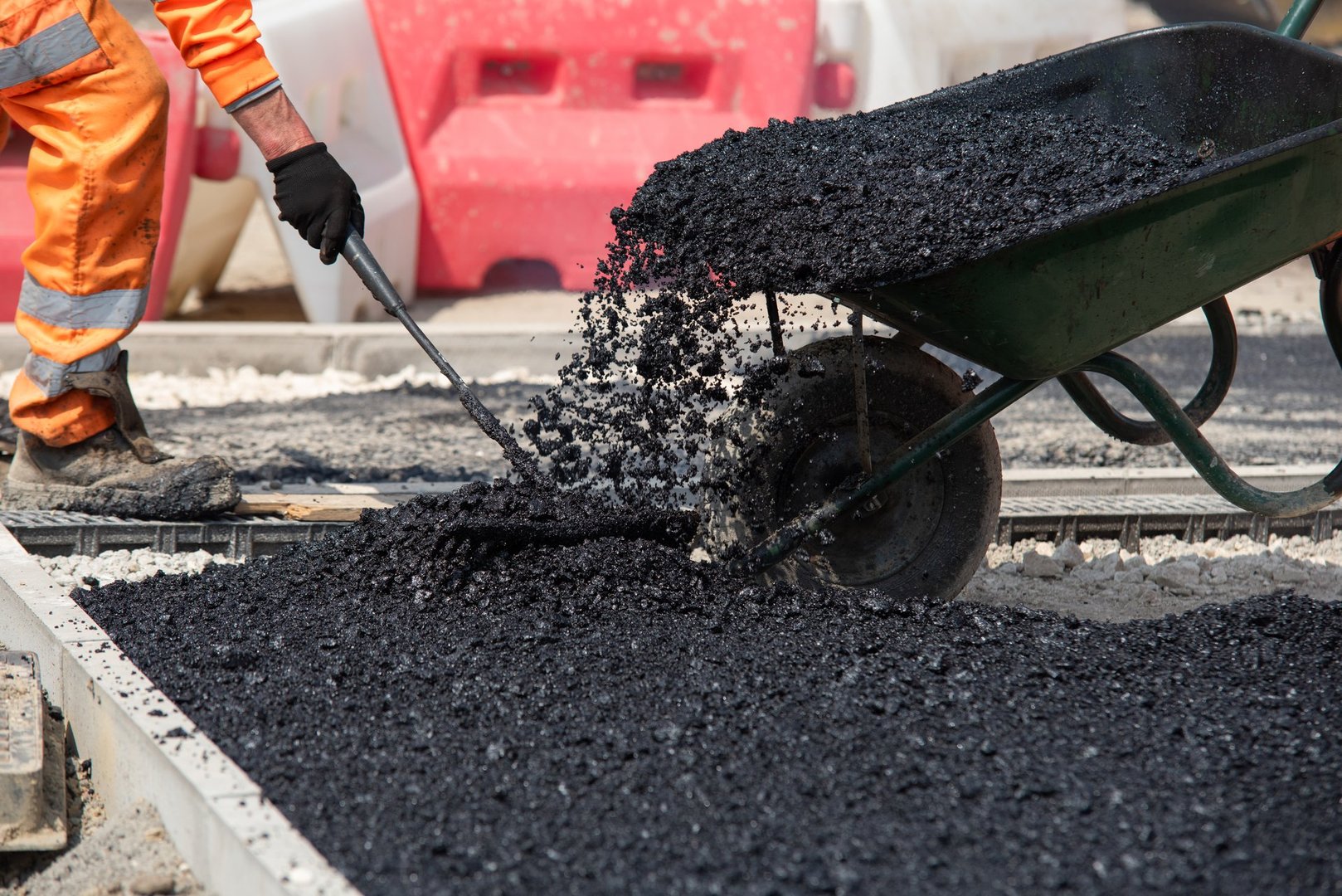 Team of road workers in bright orange uniforms repair road surface with wheelbarrow filled with hot asphalt