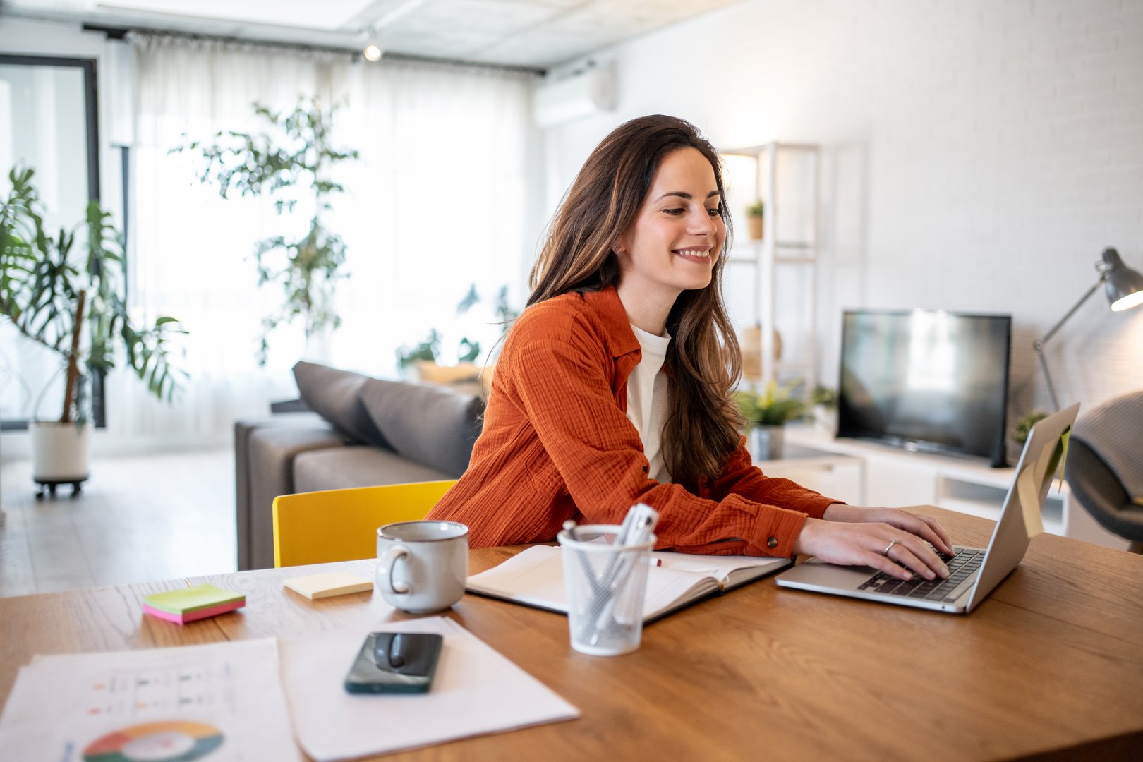 Young smiling businesswoman working from home, using laptop computer at the wooden table, with coffee cup, smartphone and notebook beside her