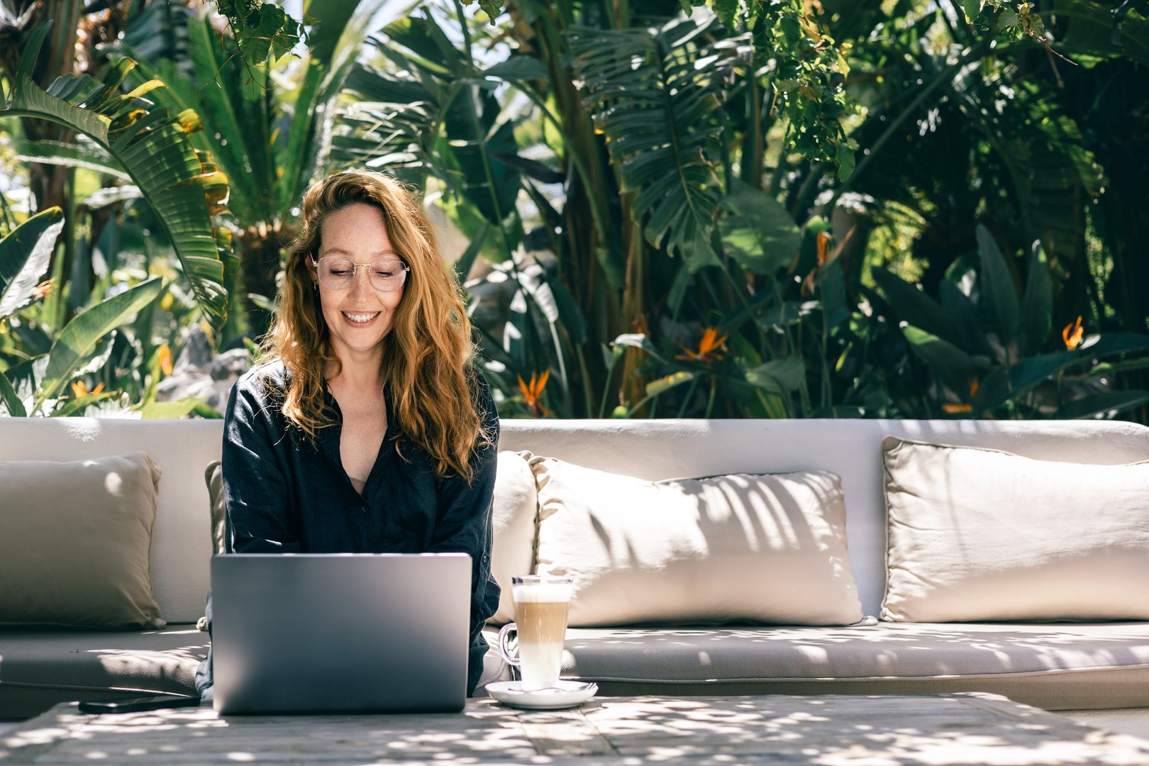 Young woman enjoying remote work in a tropical garden, sitting on a sofa with a laptop and latte. A peaceful and productive work-from-anywhere concept in a sunny and natural environment.