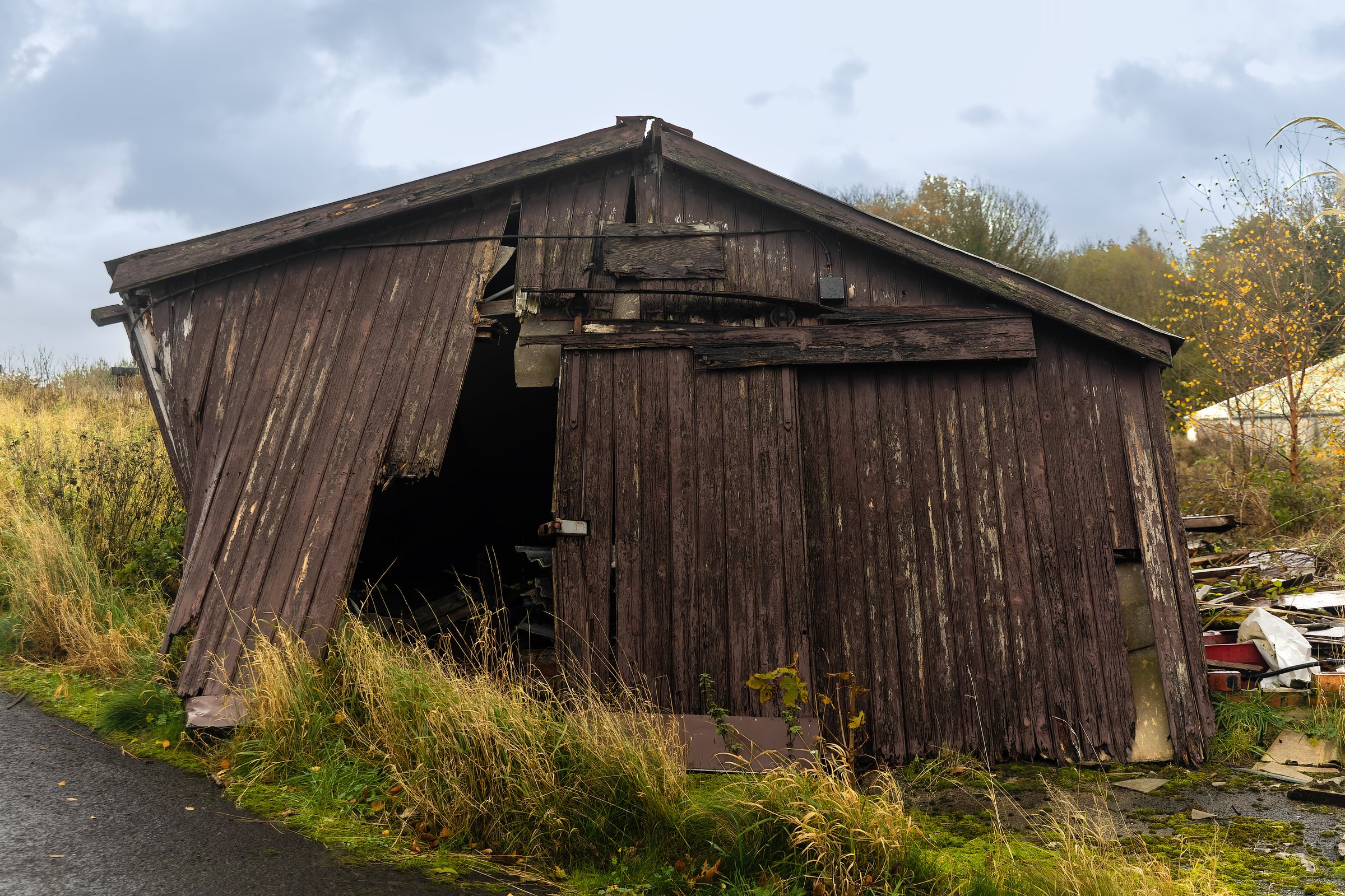 Front view of an old wooden shed with collapsing doors and leaning structure, surrounded by overgrown grass and countryside vegetation. Concept of decay, neglect, and rural abandonment
