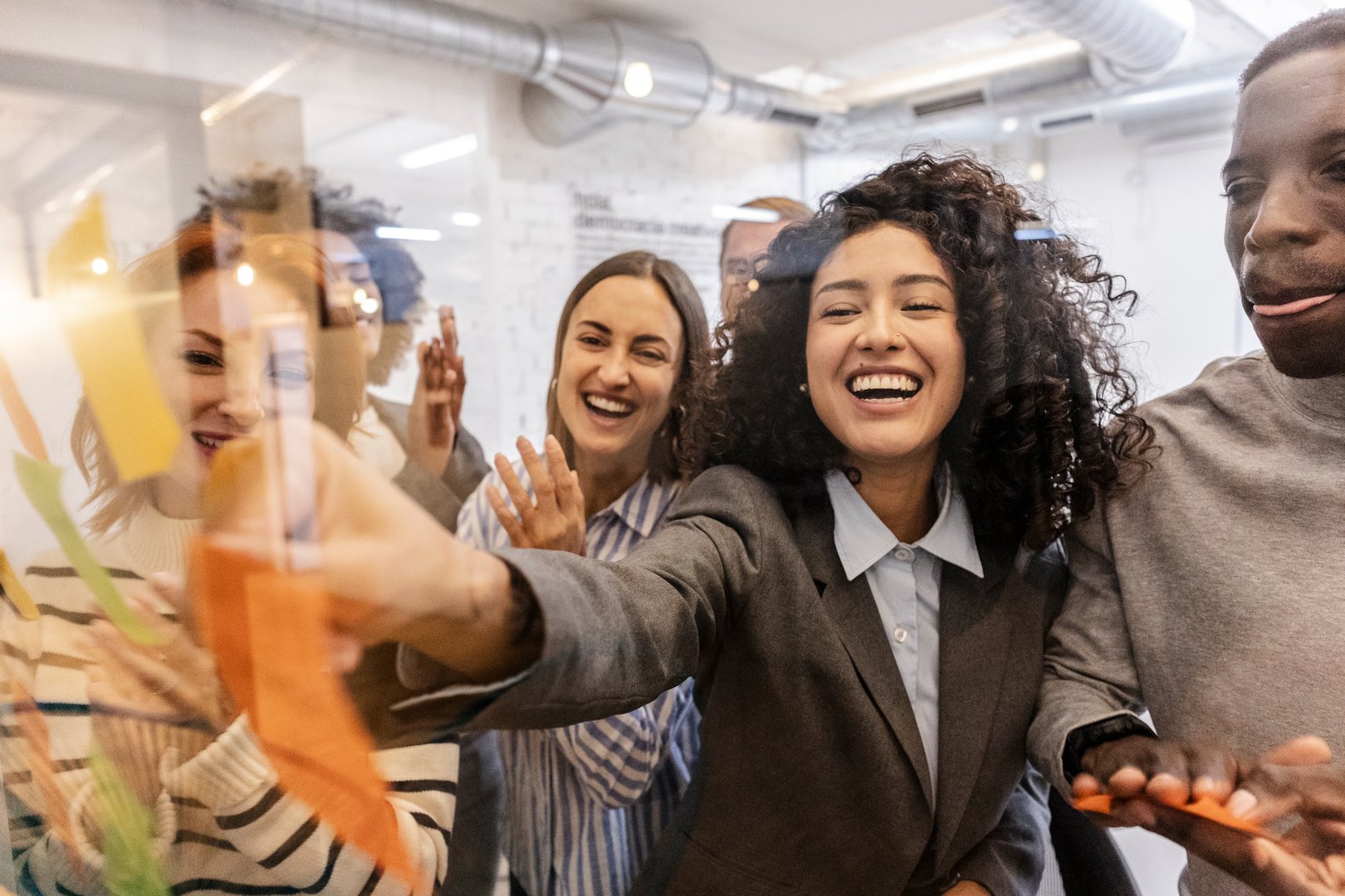 Multiracial colleagues are collaborating on a project, using colorful sticky notes on a glass wall in a modern coworking space, enjoying teamwork and celebrating diversity