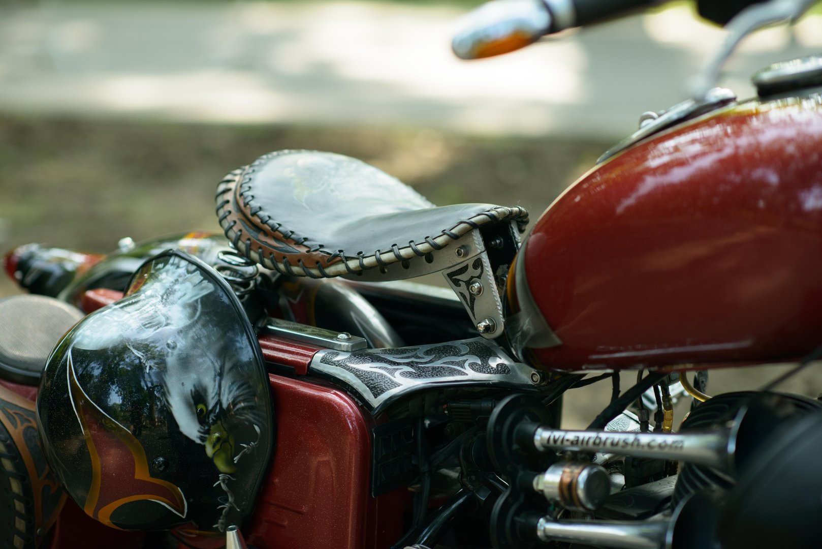Red bobber motorcycle parked in a park setting, featuring a stitched leather solo seat, engraved metal bracket, polished twin-cylinder engine and an eagle airbrushed helmet hanging beside the fuel tank.