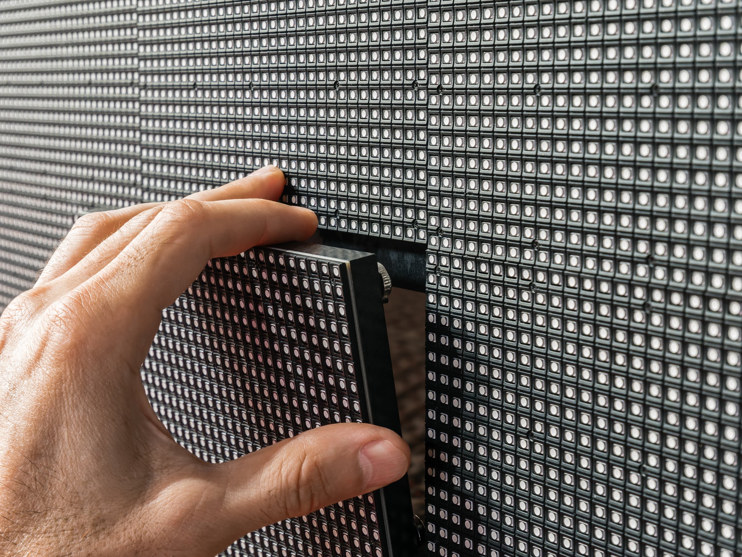 Close-up of a man's hand installing a square LED module into its place on the surface of a large LED screen.