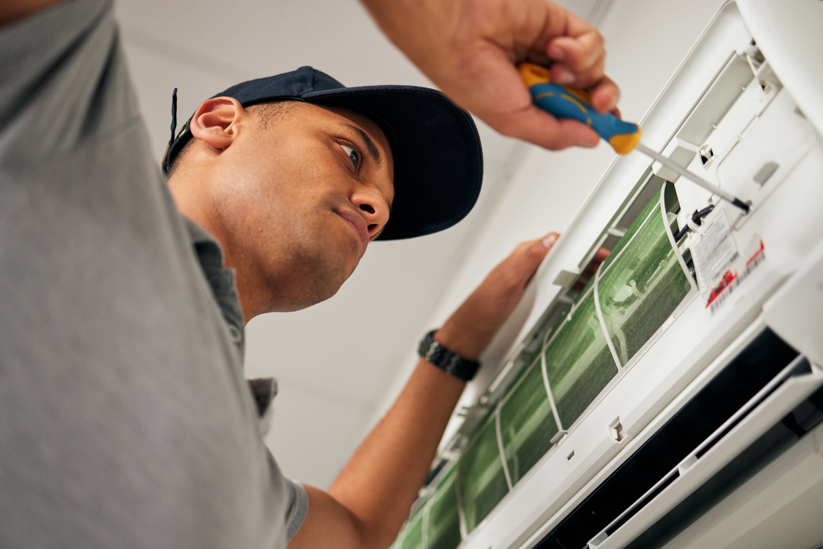 Maintenance, air conditioner and man with screwdriver for problem solving on machine from below. Aircon, ac repair and handyman service with technician, electrician or contractor work on ventilation
