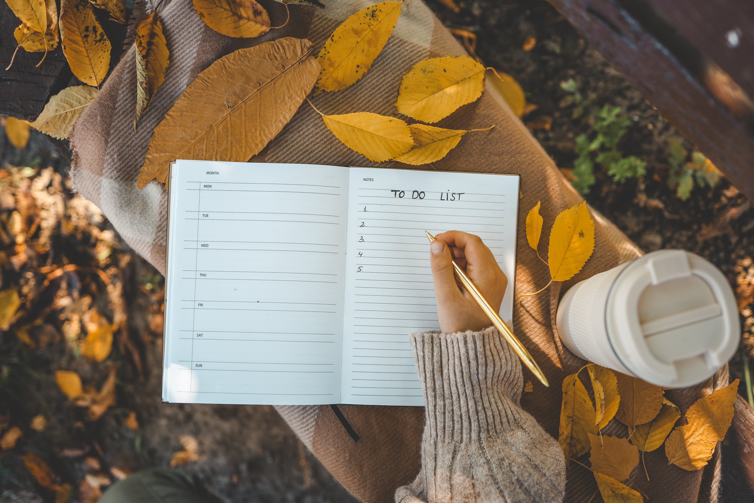 Hand writing in a planner with a gold pen, surrounded by yellow autumn leaves, a reusable cup, and cozy textures, creating a warm seasonal to-do list scene.