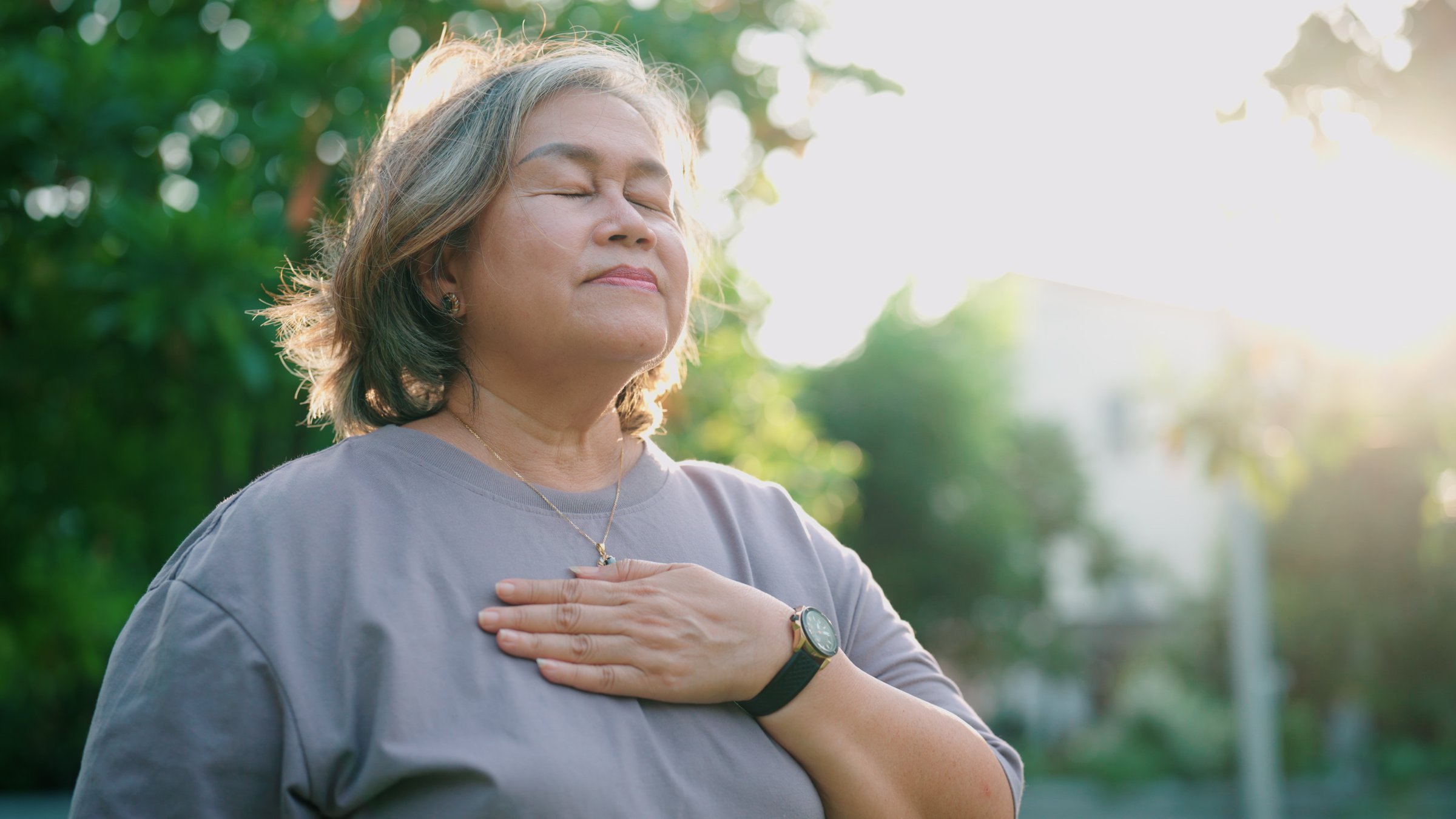 Elderly woman meditating in the park with one hand resting on her chest.