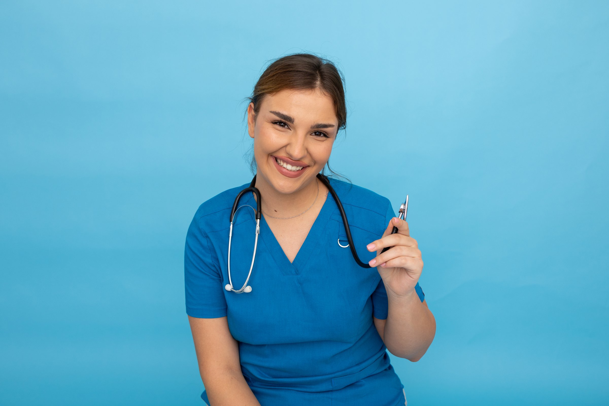 Smiling healthcare professional wearing white coat, gripping stethoscope and standing confidently against bright blue background