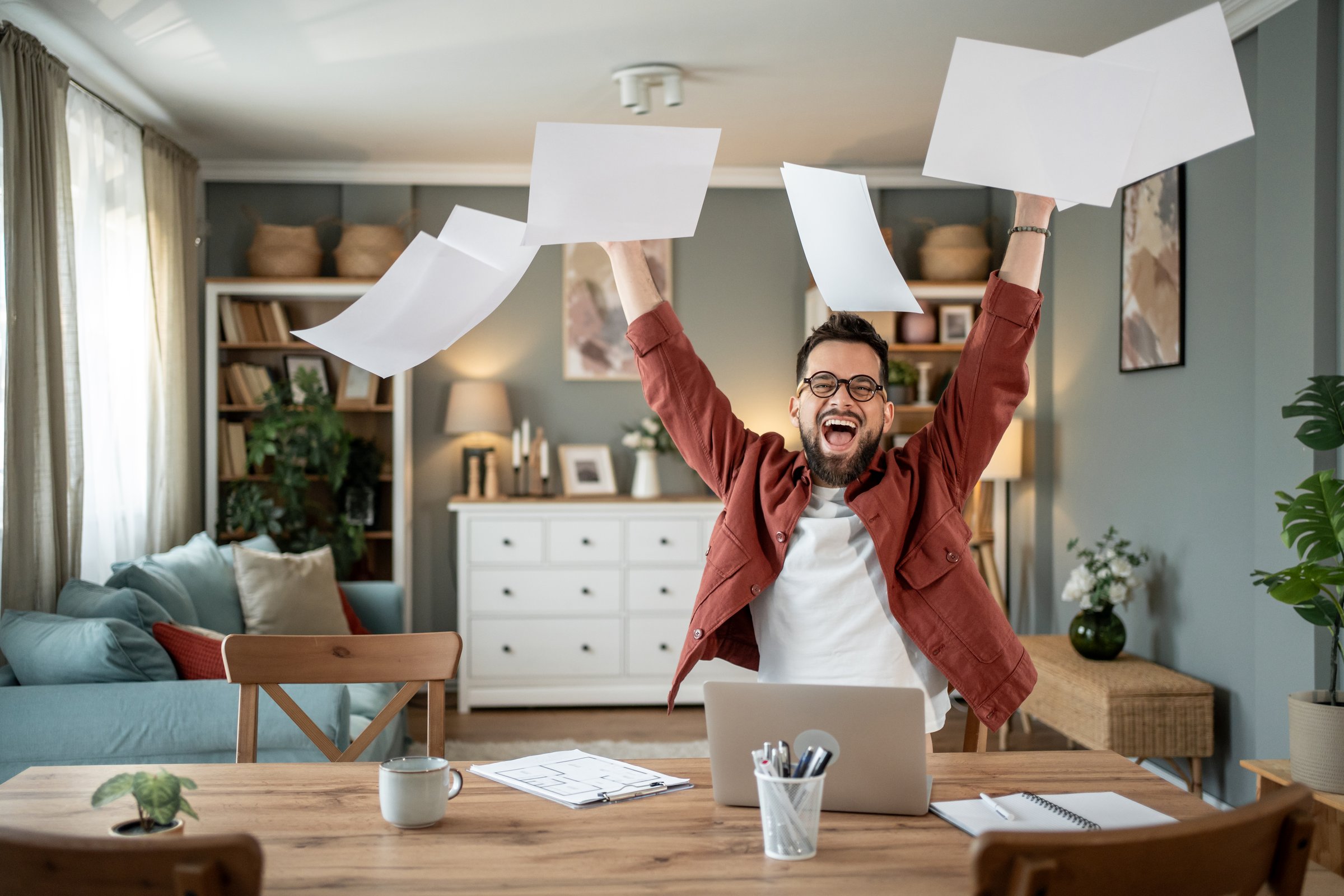 Young businessman working from home, joyfully throwing papers in the air and celebrating success while seated at his dining table, embodies the excitement of achievement and freedom